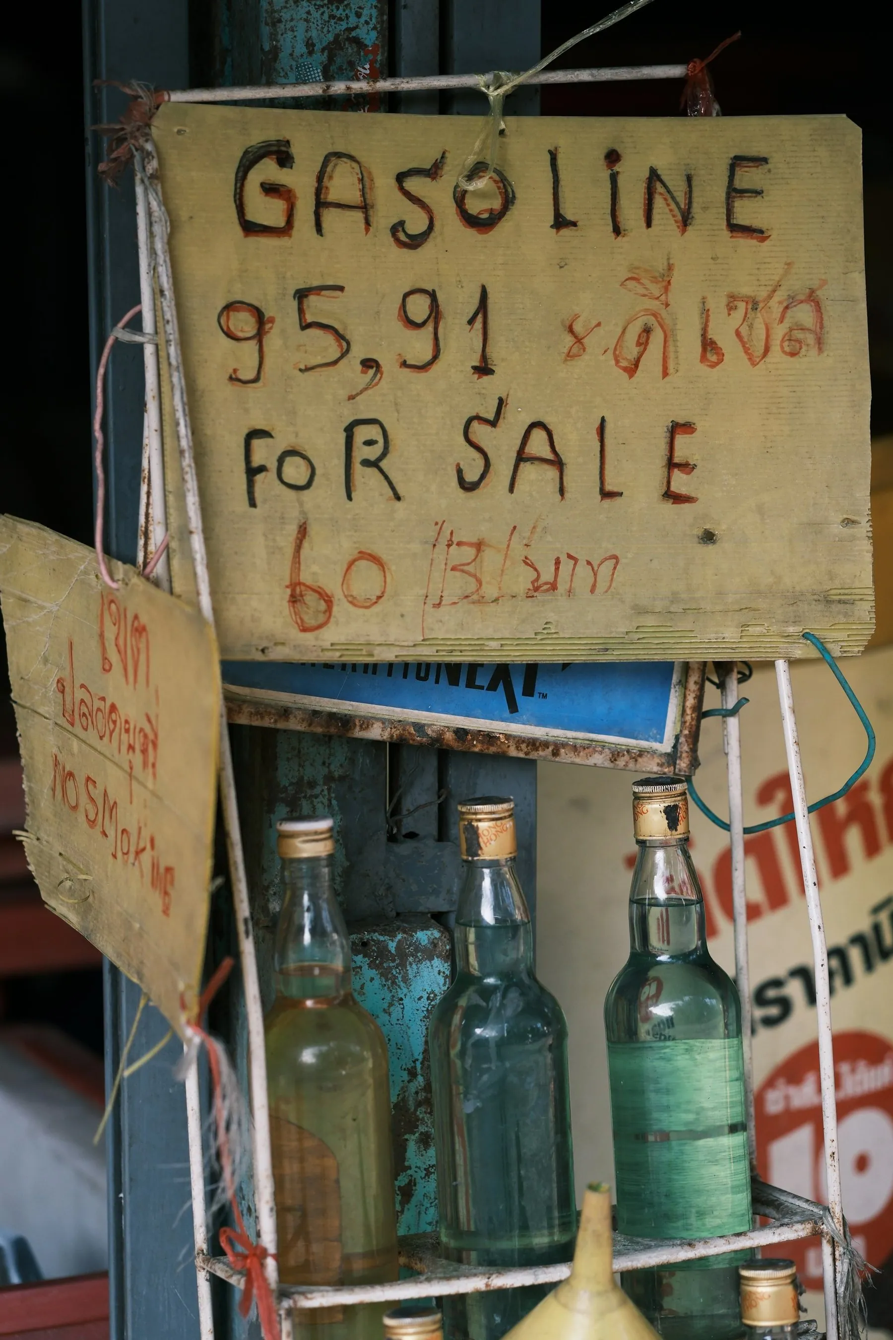 Petrol sold by the bottle at a roadside stall under a hand-lettered price sign in Thai and English