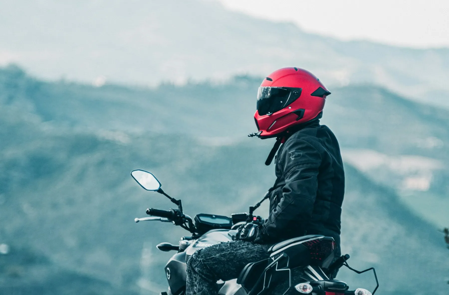 A rider in a red full-face helmet and jacket astride a large motorbike with ridgelines behind