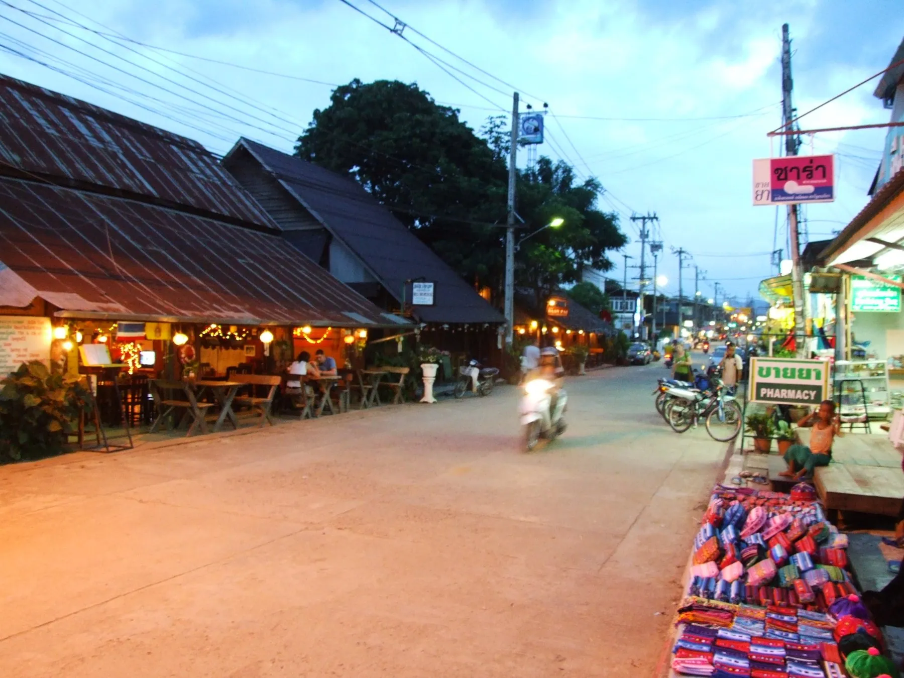 A rider passing wooden shopfronts and a pharmacy sign on the main street through Pai at dusk