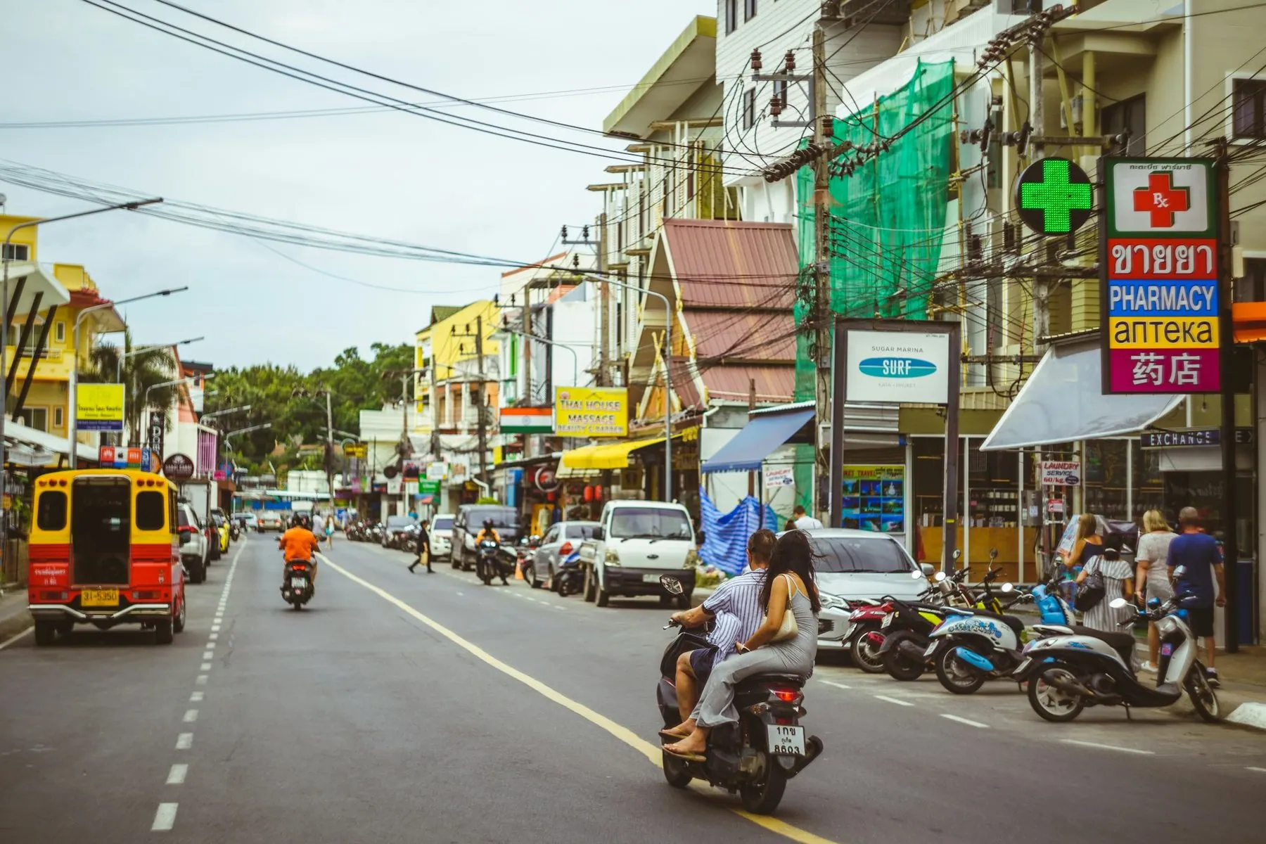Scooters and a songthaew sharing a Phuket shop street below a green pharmacy cross