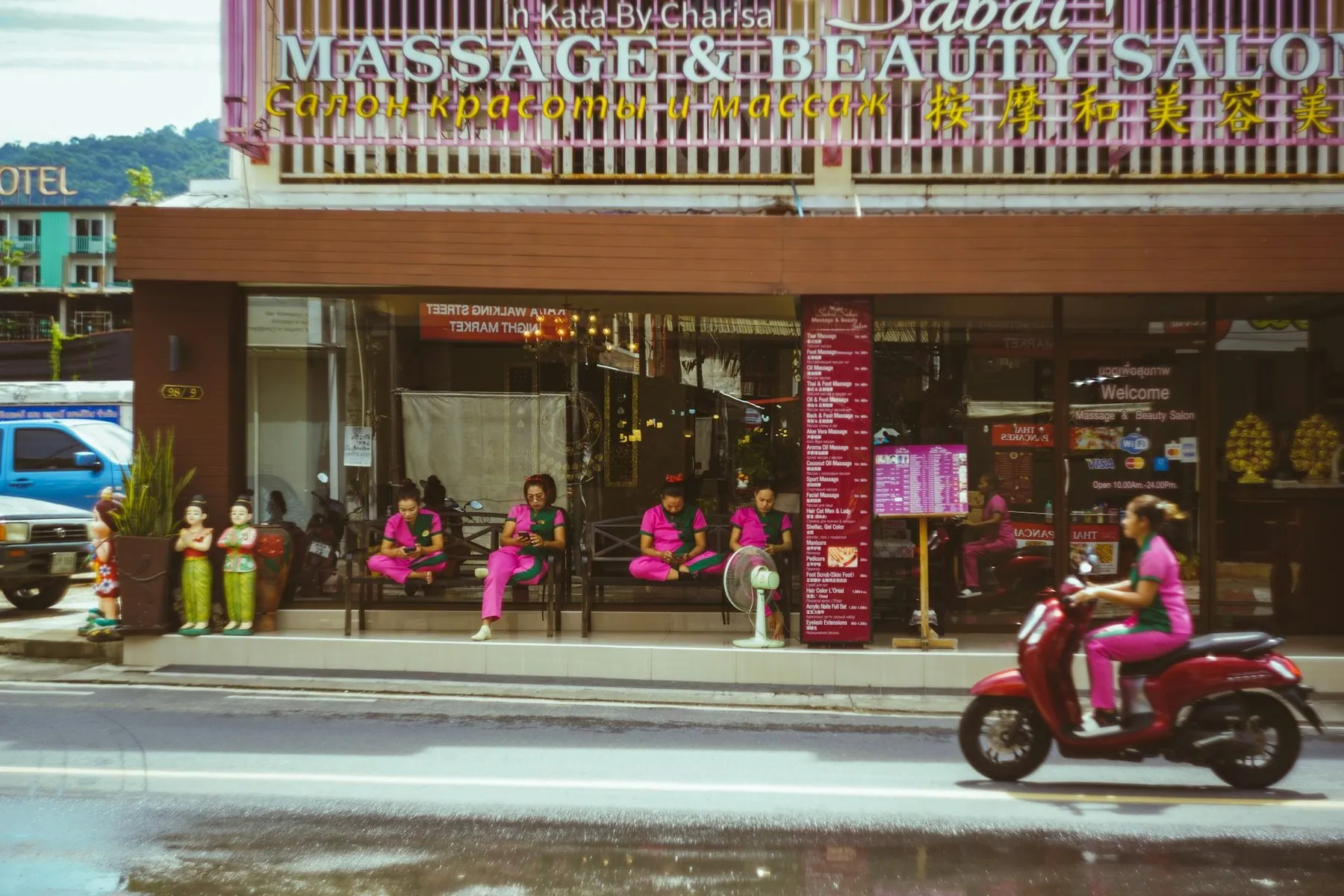 A woman riding a red scooter past a pink massage and beauty salon on the main road in Kata, Phuket