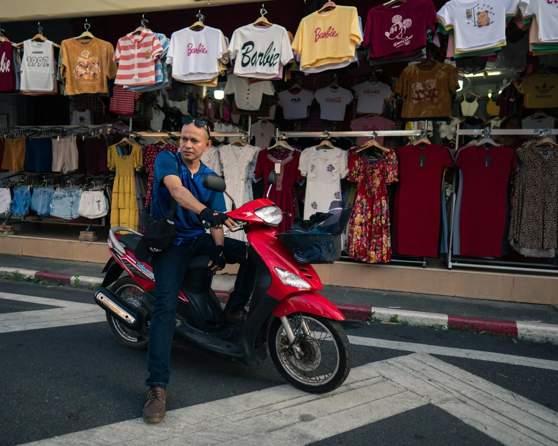 A man sitting astride a red scooter at a zebra crossing in front of a clothing stall