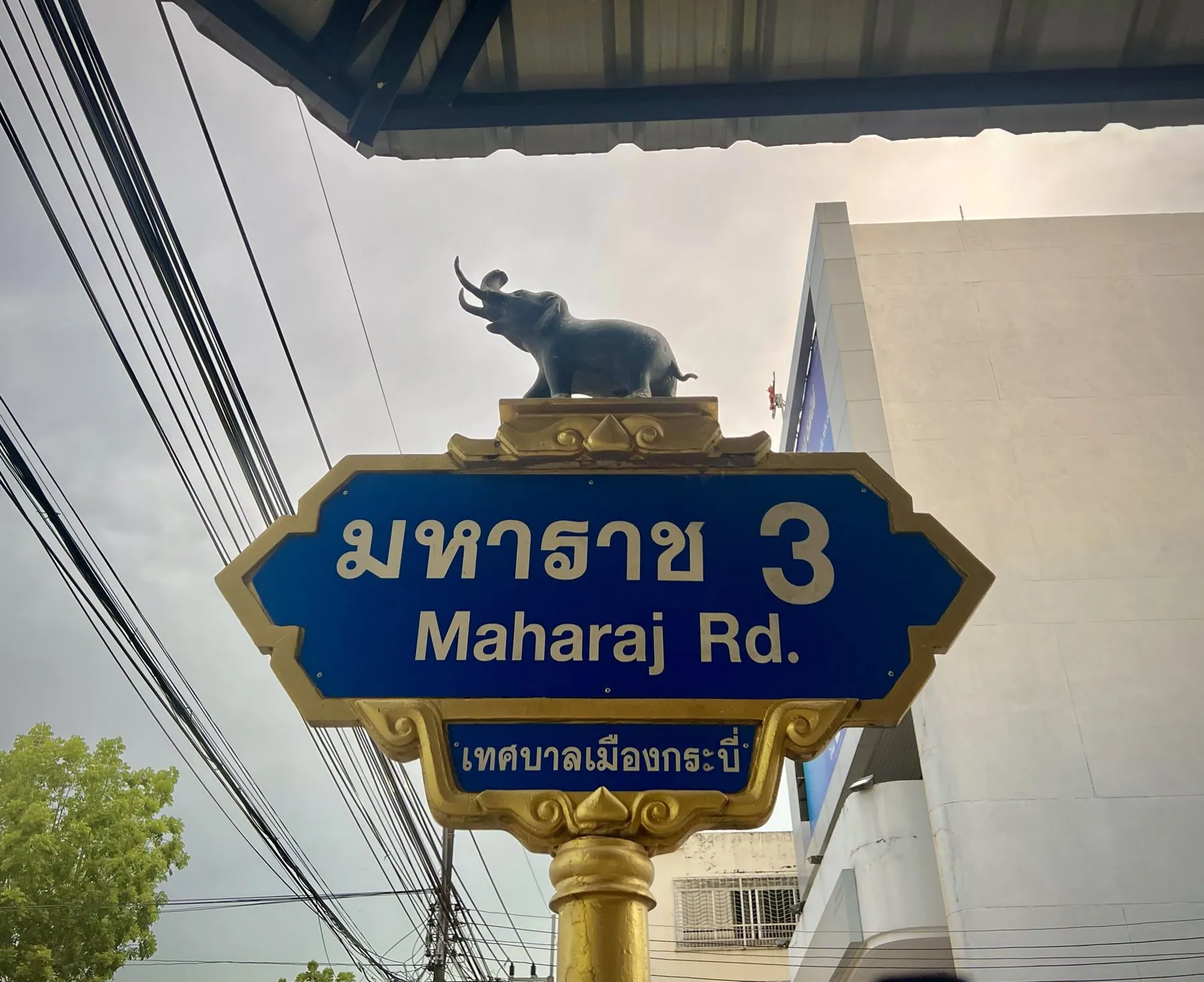 A blue and gold street sign for Maharaj Road in Krabi town, topped by a small elephant figure