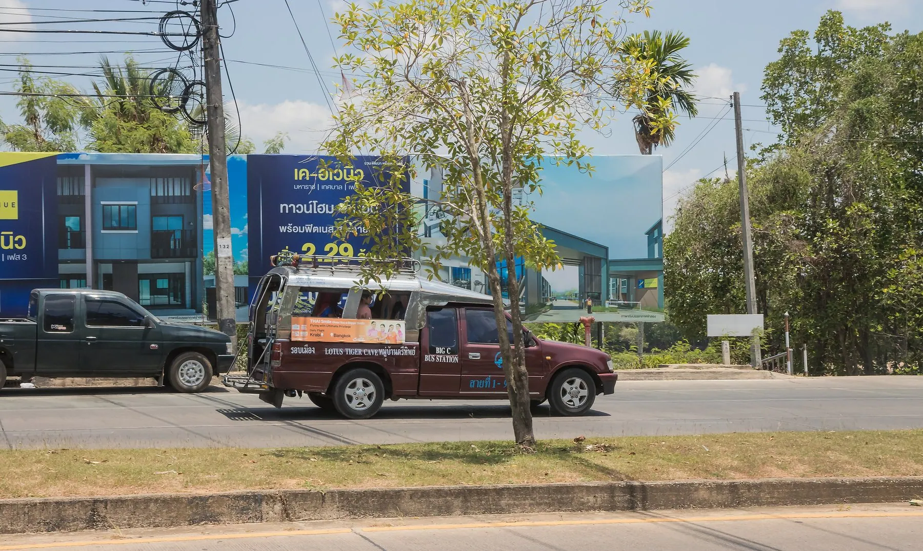 A red songthaew pulling away on a Krabi road with limestone cliffs rising behind the roofline