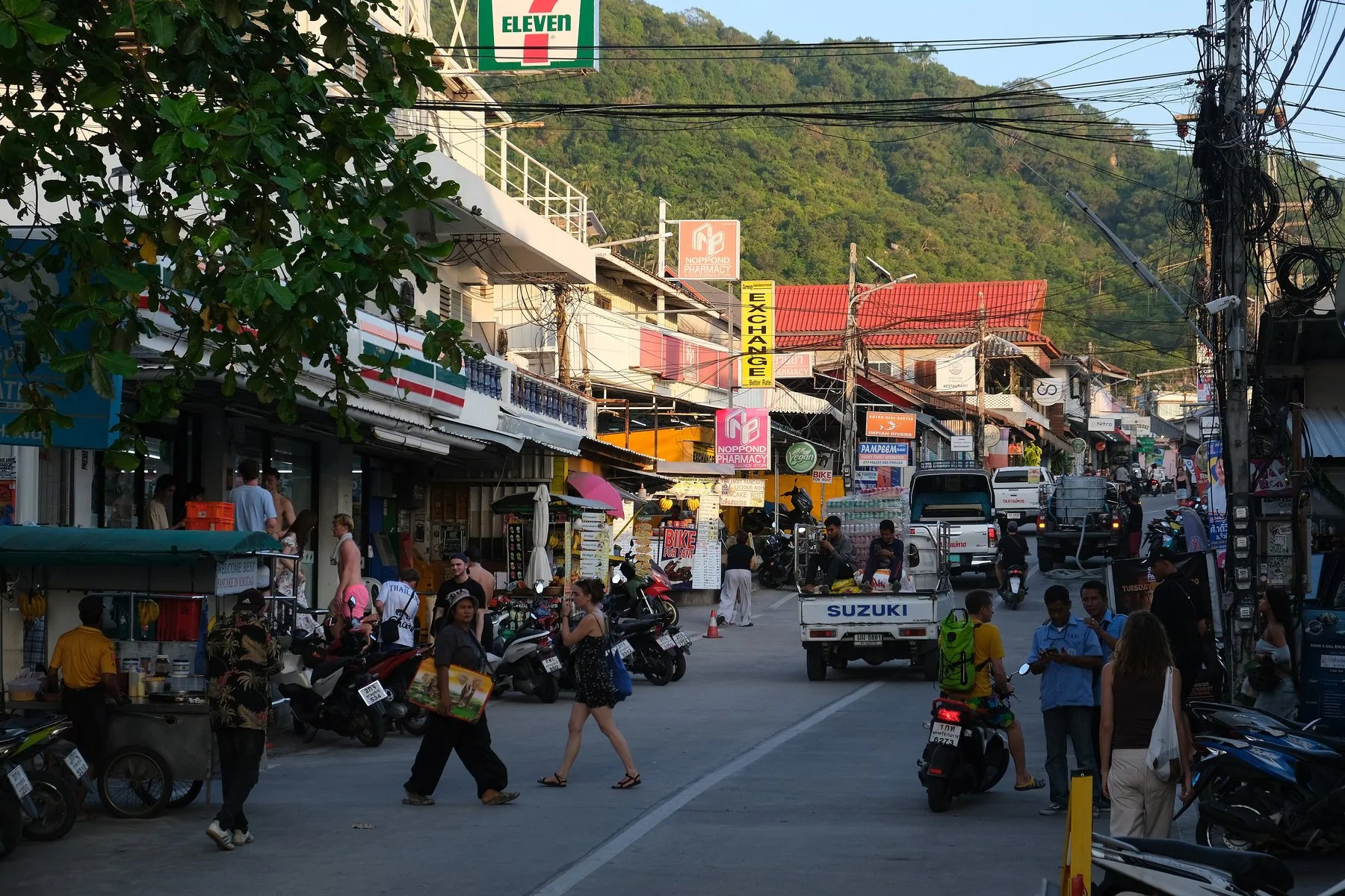 The main street in Mae Haad on Koh Tao, scooters and pickups outside a 7-Eleven below wooded hills