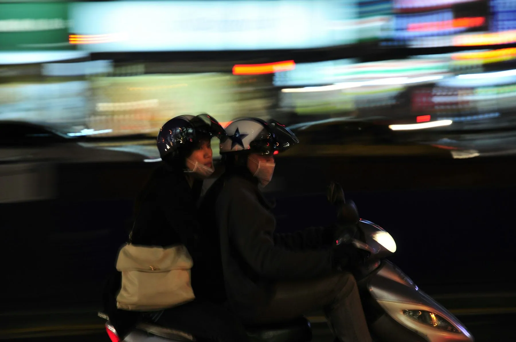 Two helmeted riders sharing an automatic scooter through blurred city lights at night