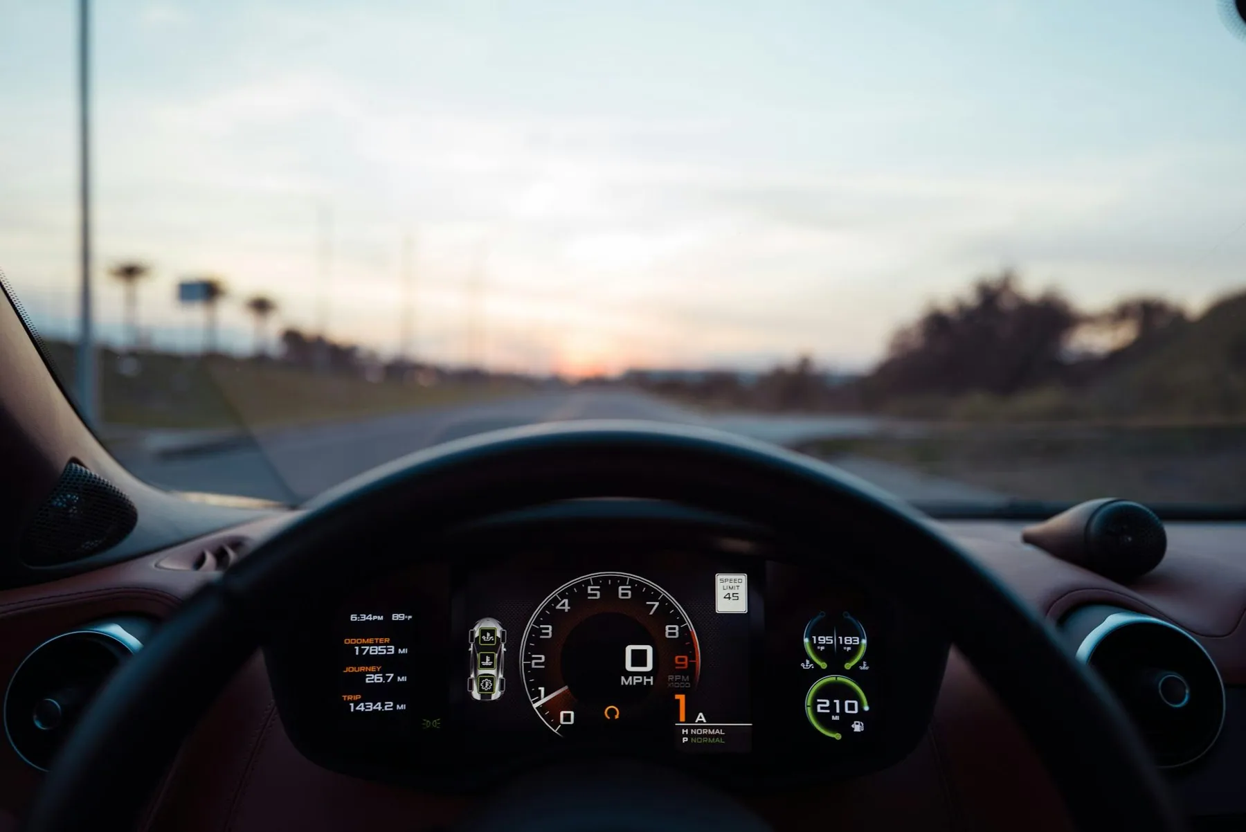 A car's lit instrument cluster and steering wheel seen from the driver's seat, with an open road and a dusk sky through the windscreen