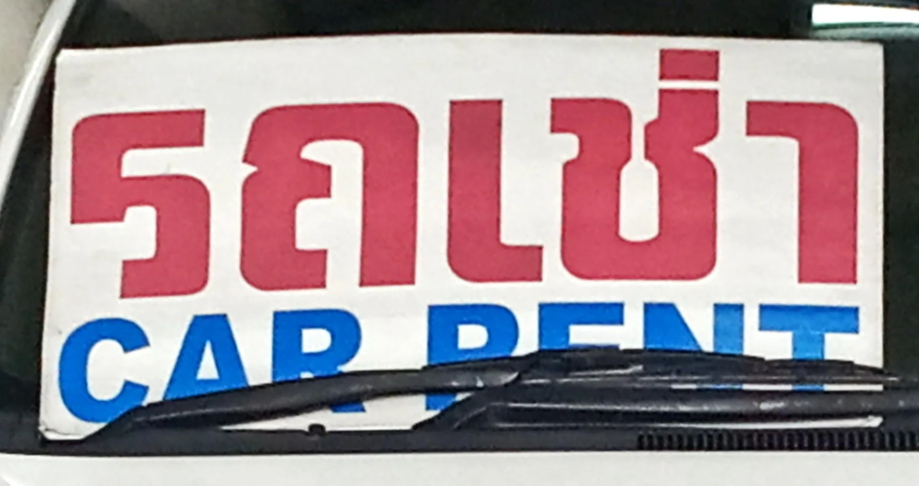 A printed sign reading รถเช่า and CAR RENT propped inside a car windscreen behind the wiper blade
