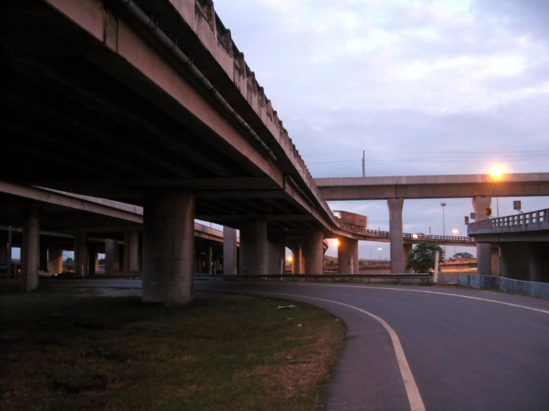A slip road curving beneath the concrete viaducts of Bangkok's outer ring road at dusk