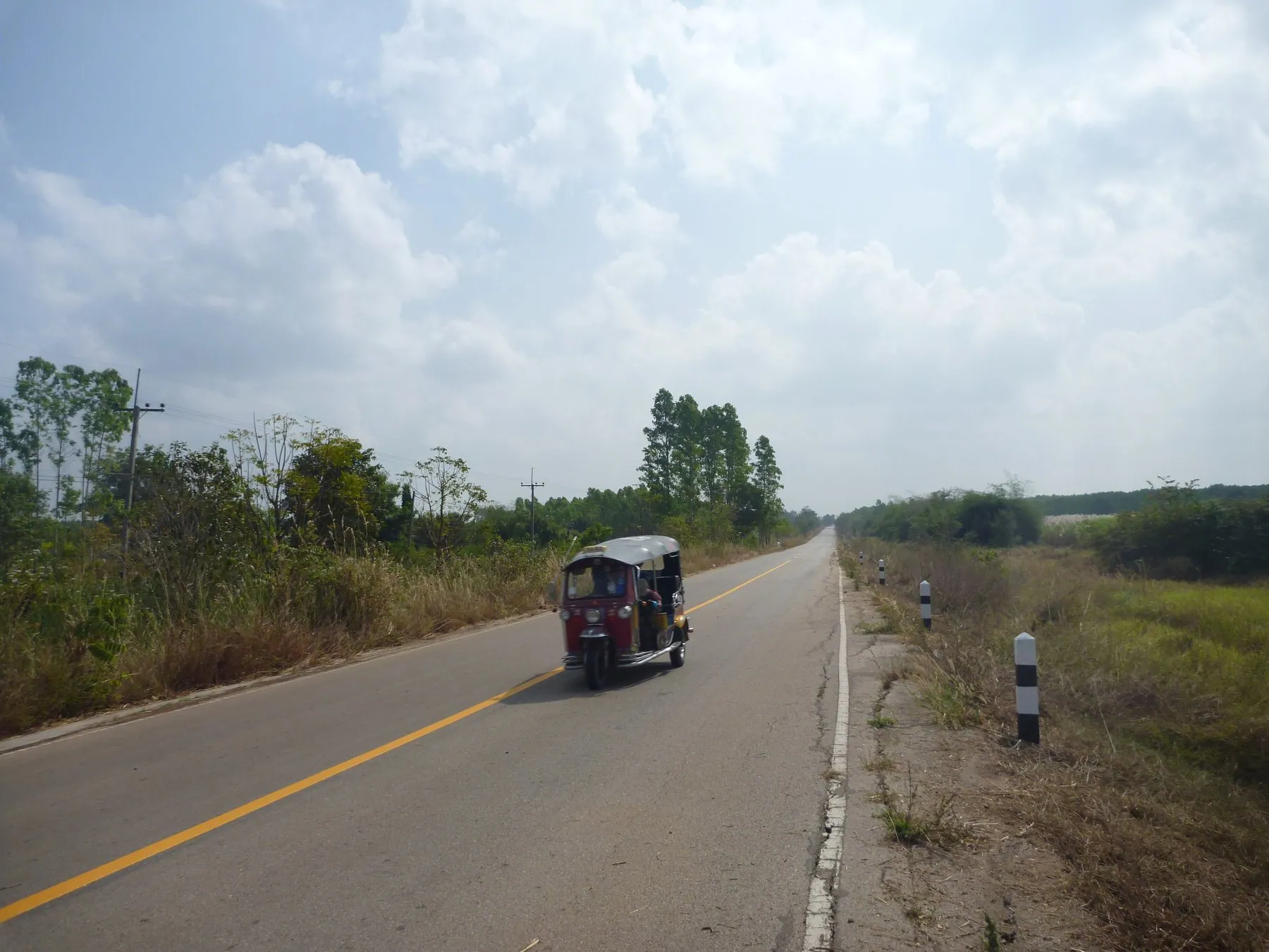 A tuk-tuk on an empty straight road through flat country in Sa Kaeo province on the way to the Cambodian border