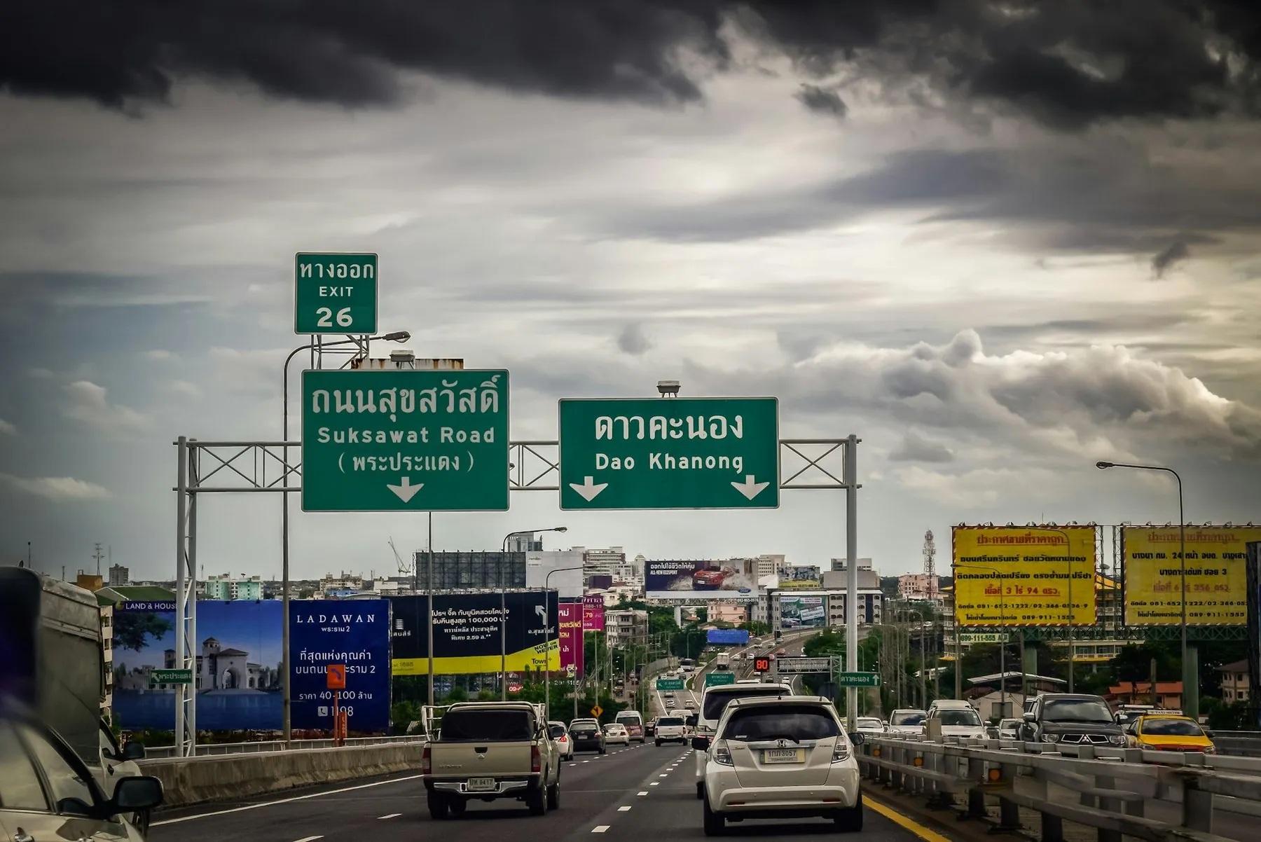 Traffic on a Bangkok expressway under heavy cloud, green overhead gantry signs in Thai and English