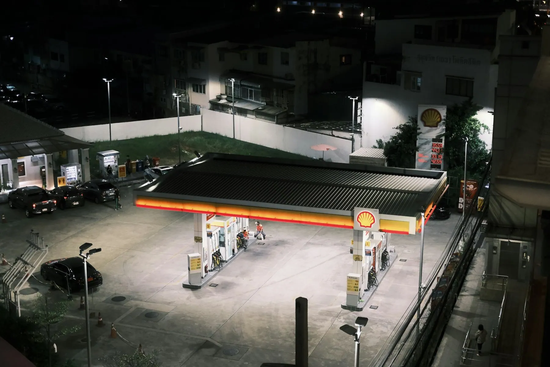 A lit petrol station forecourt in Bangkok at night with cars and motorbikes waiting at the pumps