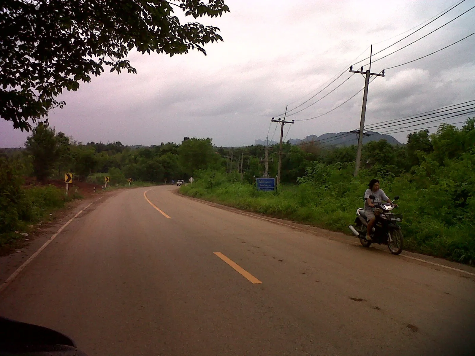 An empty two-lane road running north out of a small Isaan town, a single motorbike in the distance