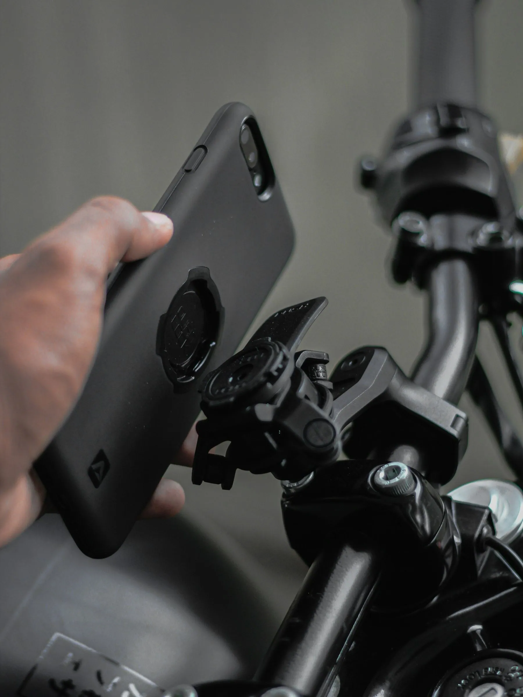 A hand holding a phone with a mounting puck on the back, about to click it into a clamp on a set of handlebars
