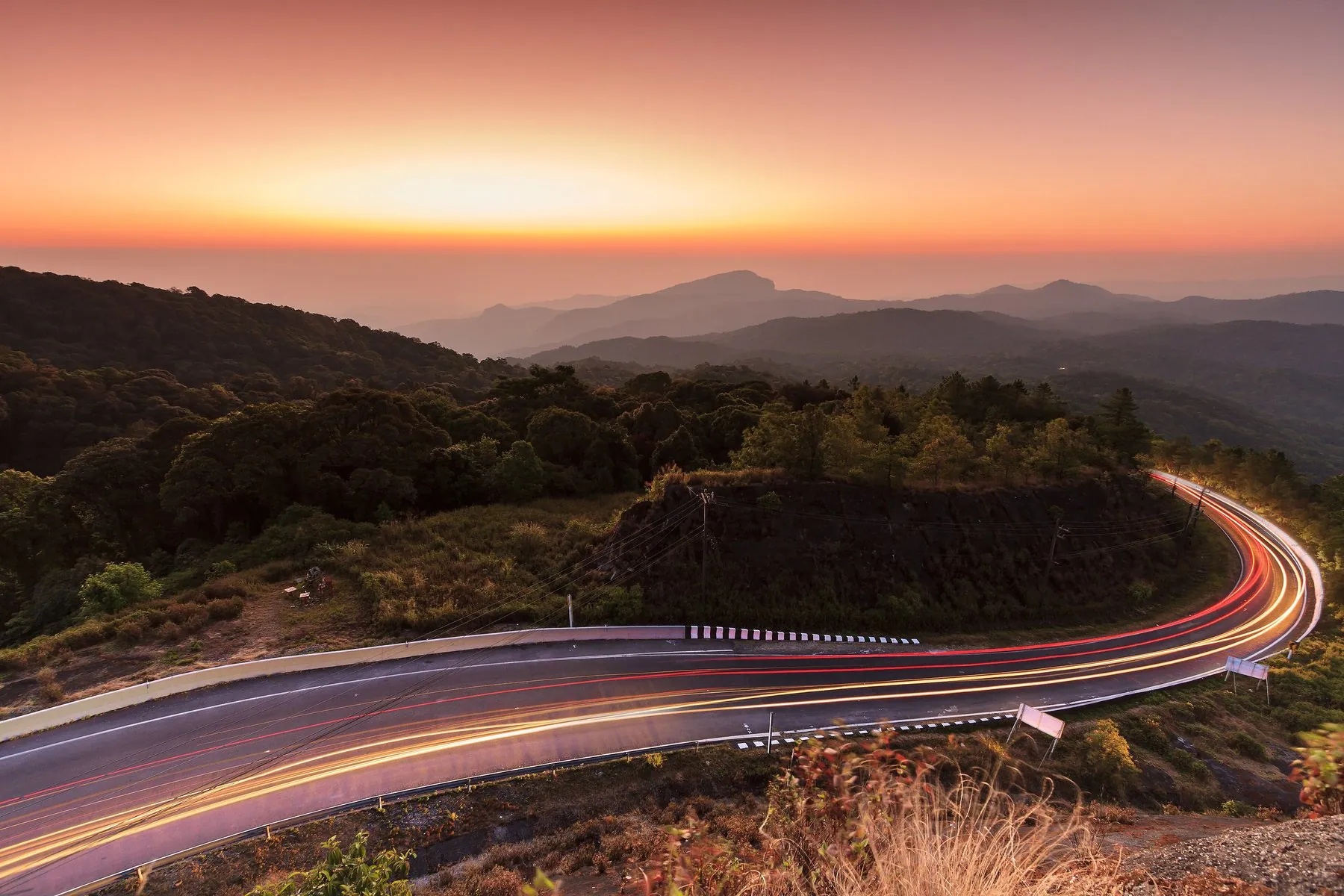 Headlight and tail-light trails curving along a mountain road at sunrise, ridges stacked into the distance