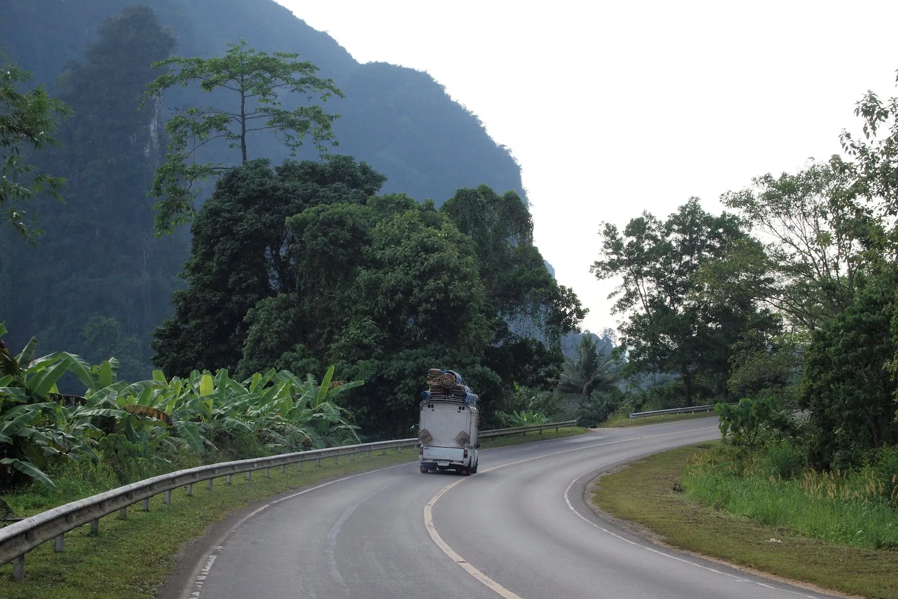 An almost empty road through rainforest with karst peaks ahead, a single lorry in the distance