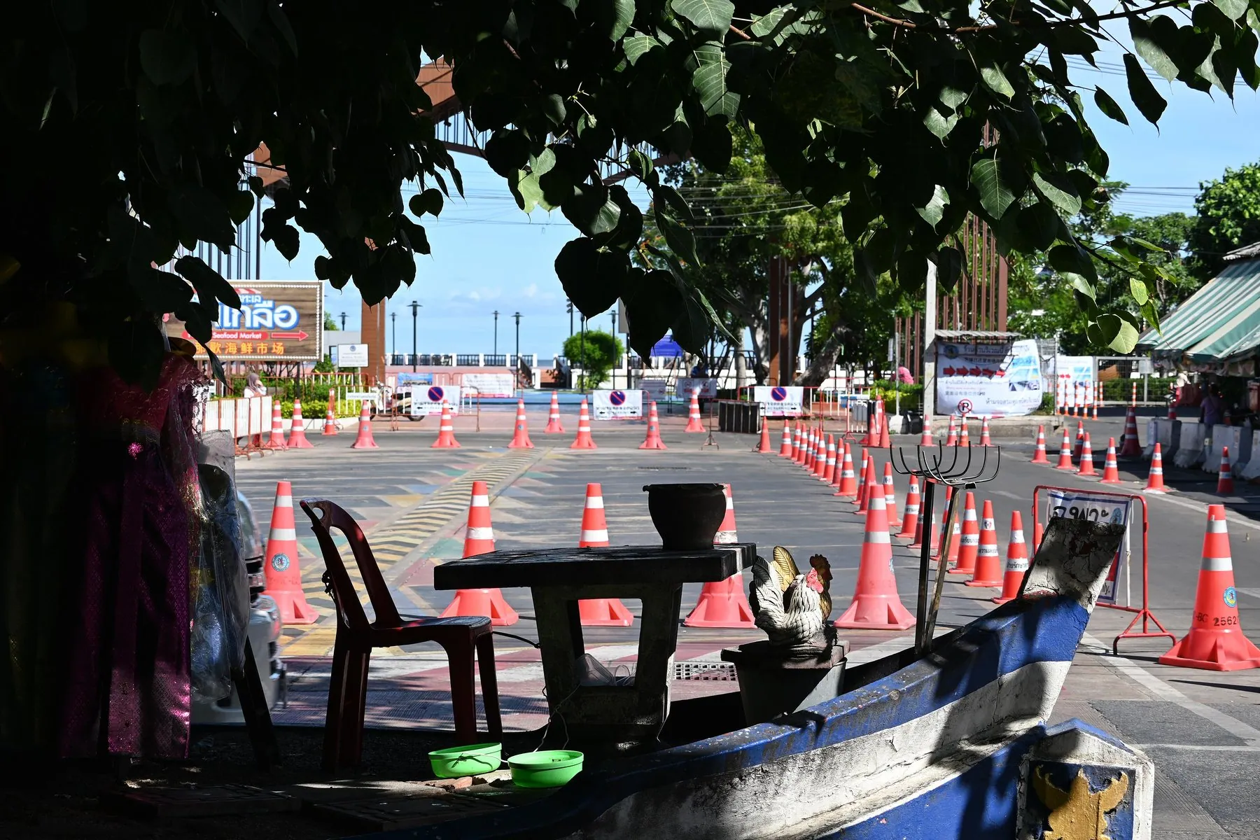 A line of orange cones and barriers narrowing a Thai road, seen from a shaded roadside stall