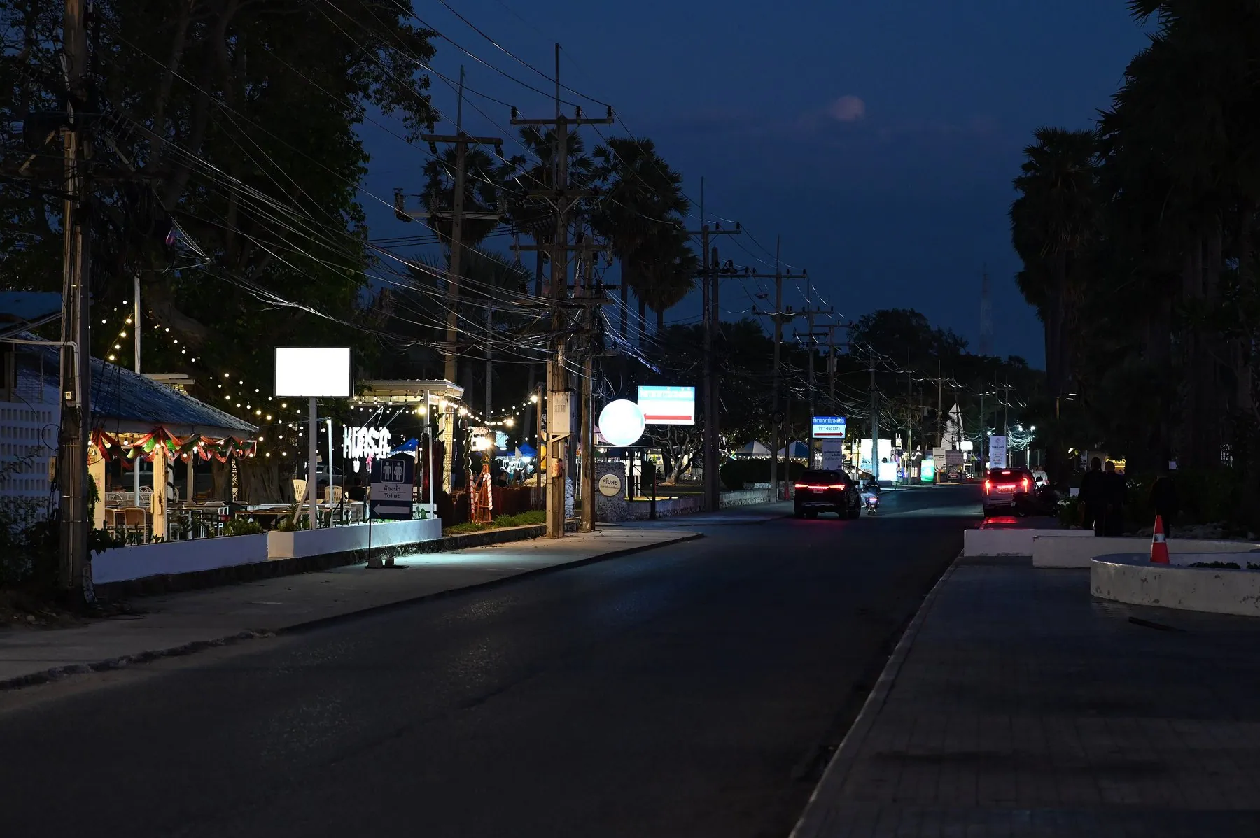 A quiet two-lane road at dusk under a deep blue sky, a pickup and a motorbike running with their lights on past shophouses strung with lights