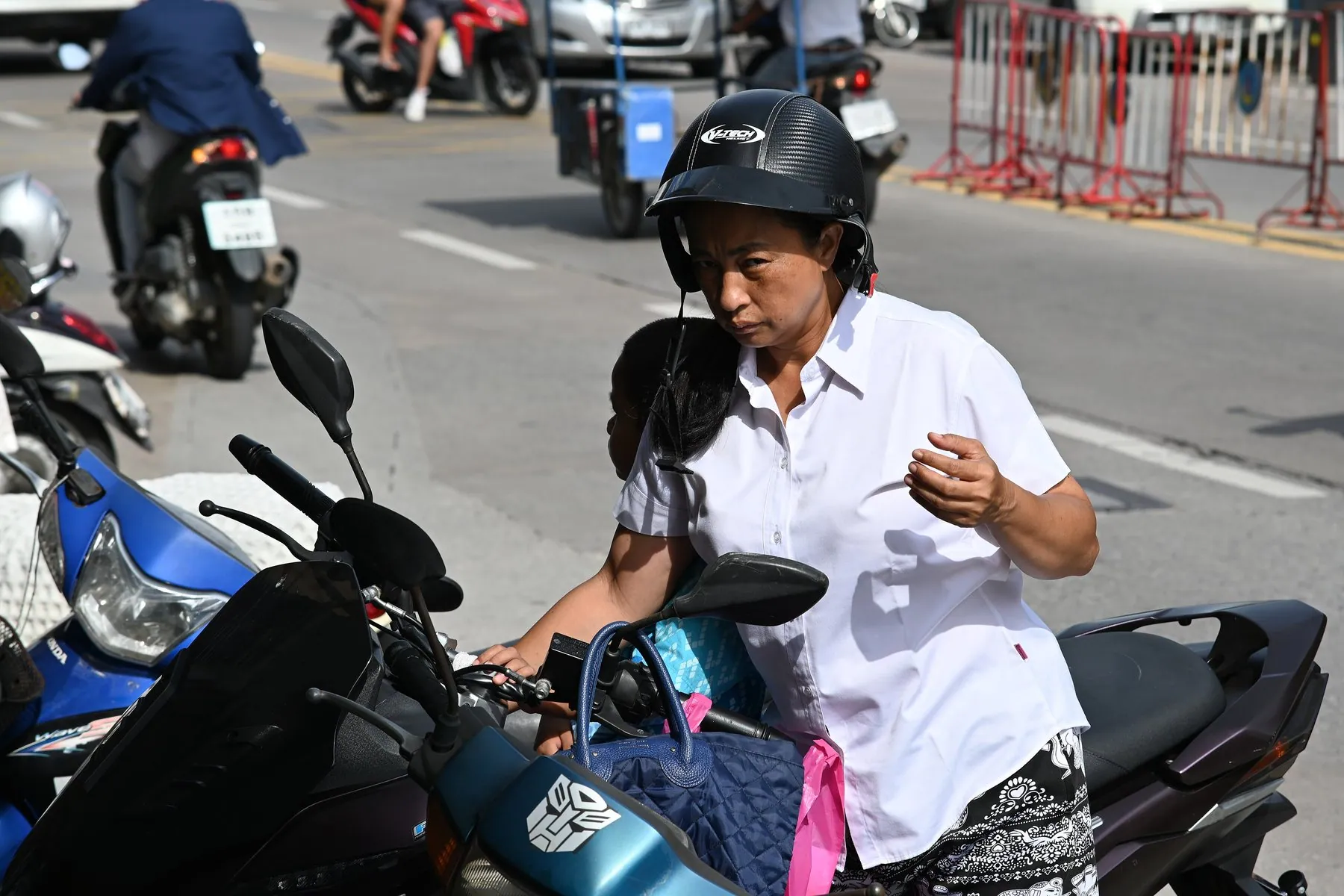 A helmeted rider strapping a load to a parked scooter on a Thai city street