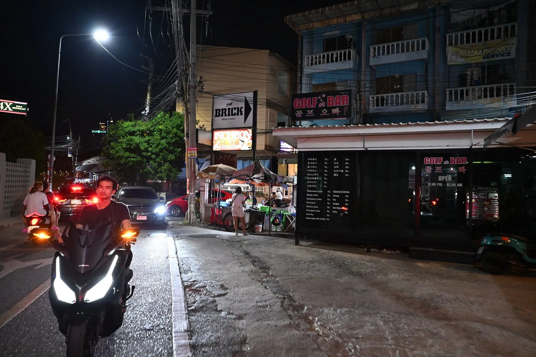 A scooter ridden towards the camera on a wet street at night with its headlight and both amber front indicators lit, a car's tail lights behind
