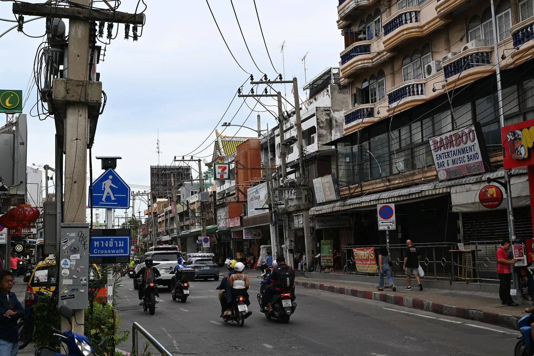 Motorcyclists riding along the near side of a Thai shopping street beneath tangled overhead power lines