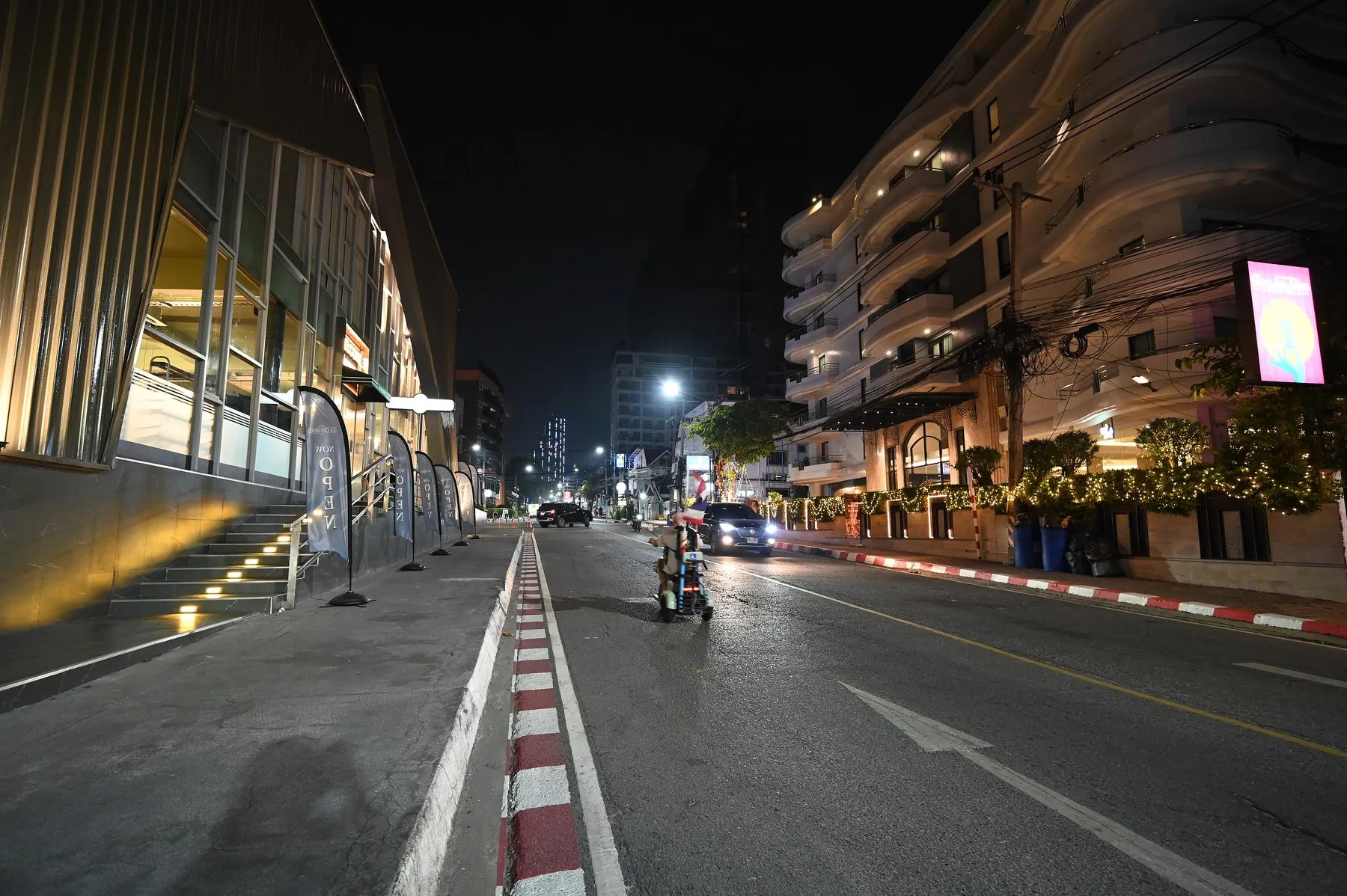 A lone motorcyclist riding down the middle of an empty Thai street at night between lit shopfronts