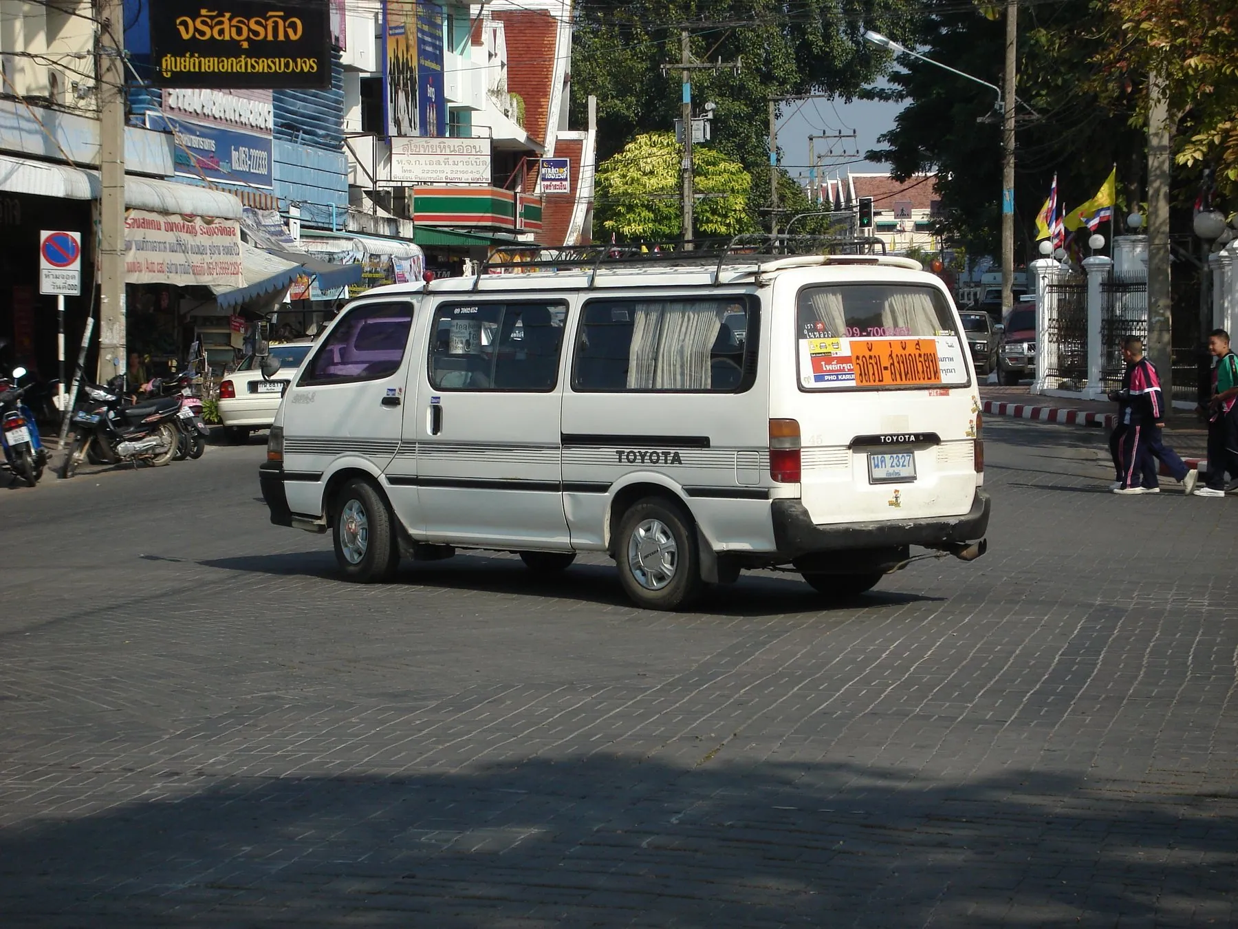 A white Toyota Hiace school van with a roof rack and Thai lettering across its tailgate turning through a town junction, two pupils walking on the pavement behind