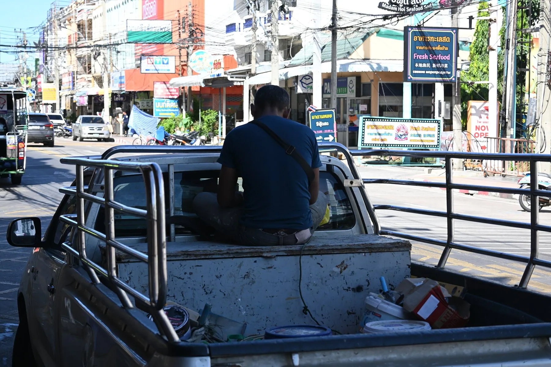 Two passengers sitting in the open bed of a pickup truck as it drives along a Thai shopping street