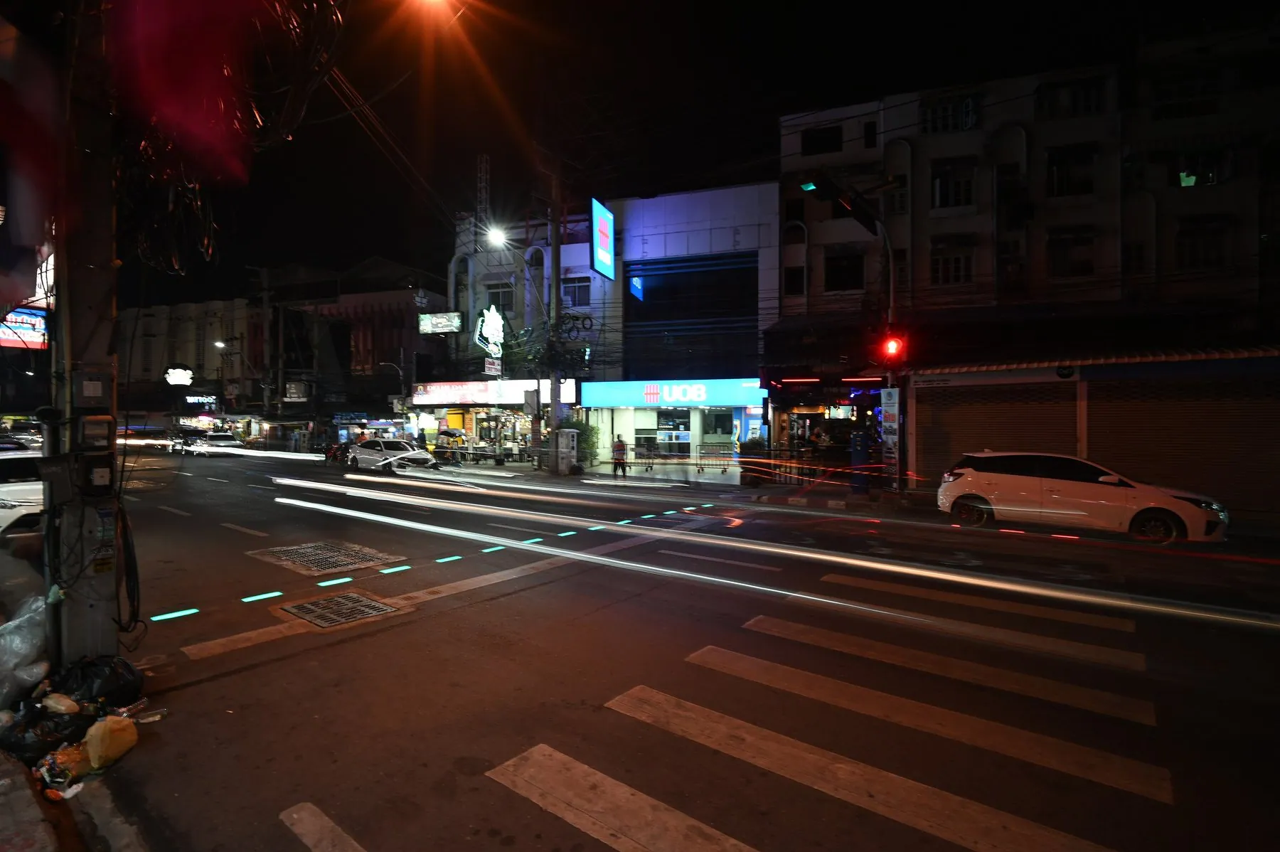 White zebra-crossing bars painted across a Thai city junction with headlight trails behind them
