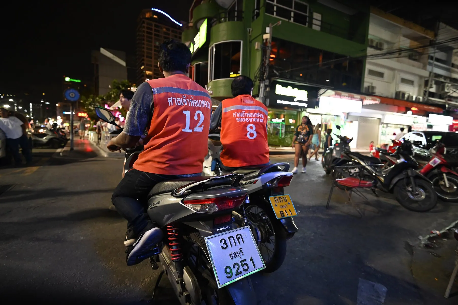 Two motorcycle-taxi riders in numbered orange vests waiting at a Thai city junction after dark