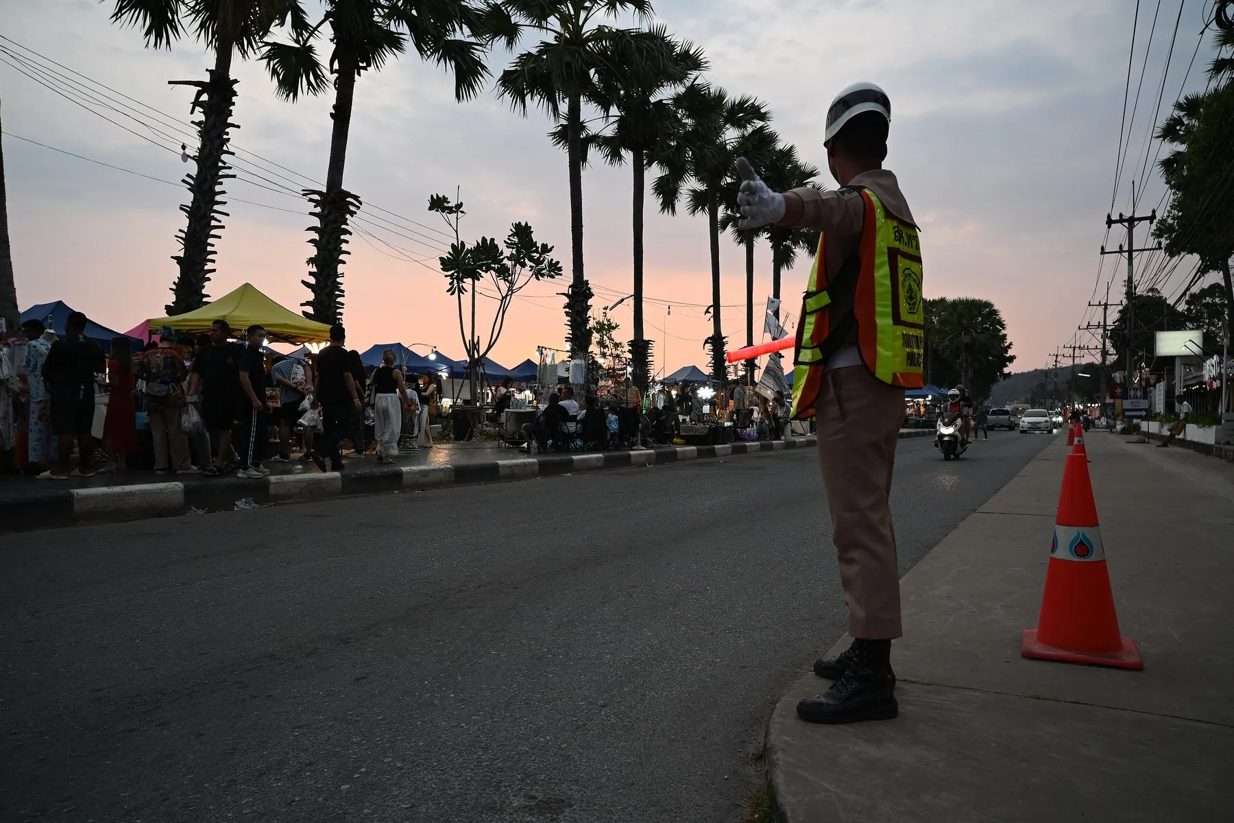 A Thai traffic officer in a reflective jacket waving vehicles through a palm-lined street at dusk