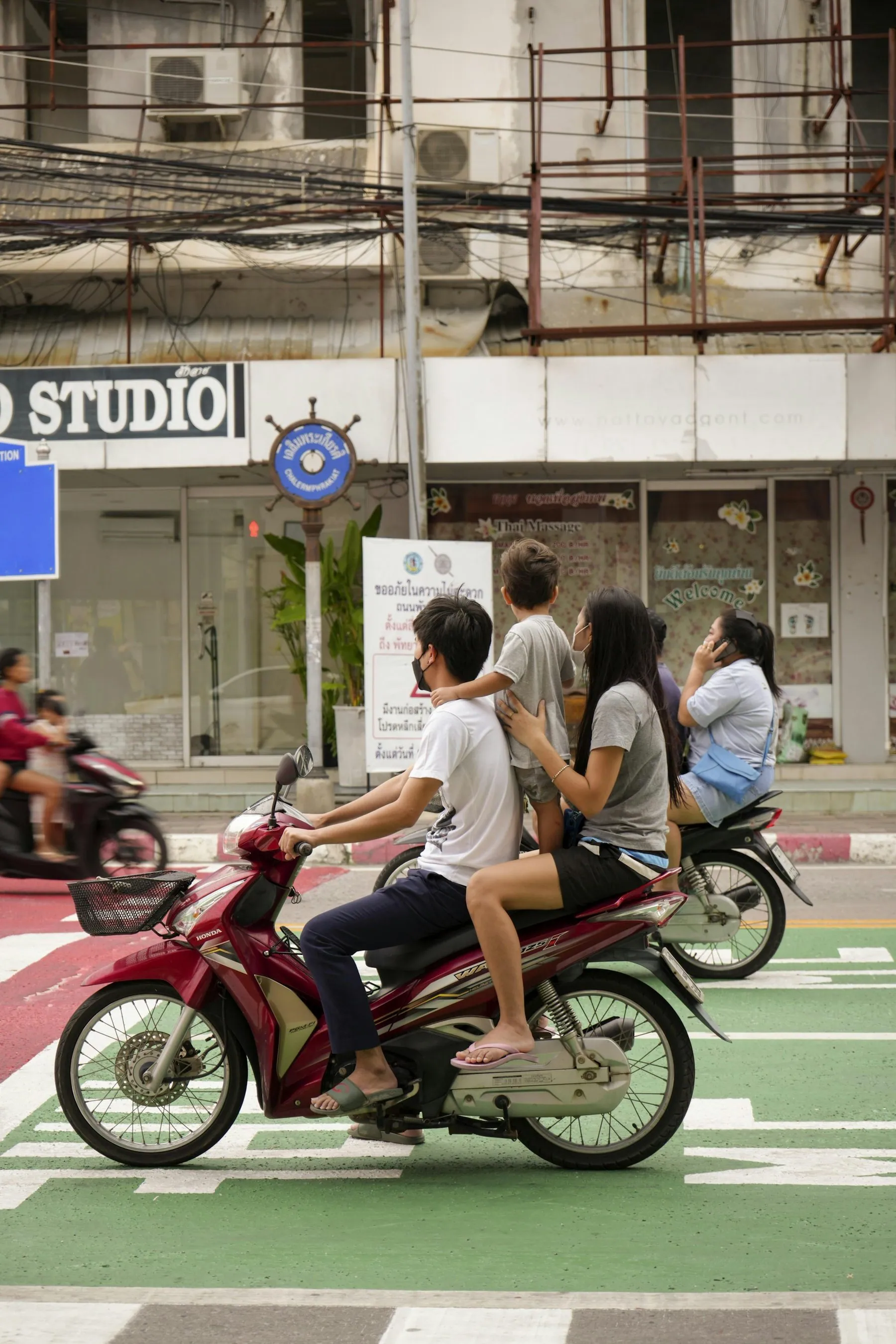 Three young people riding bare-headed on a single scooter across a marked crossing in Pattaya