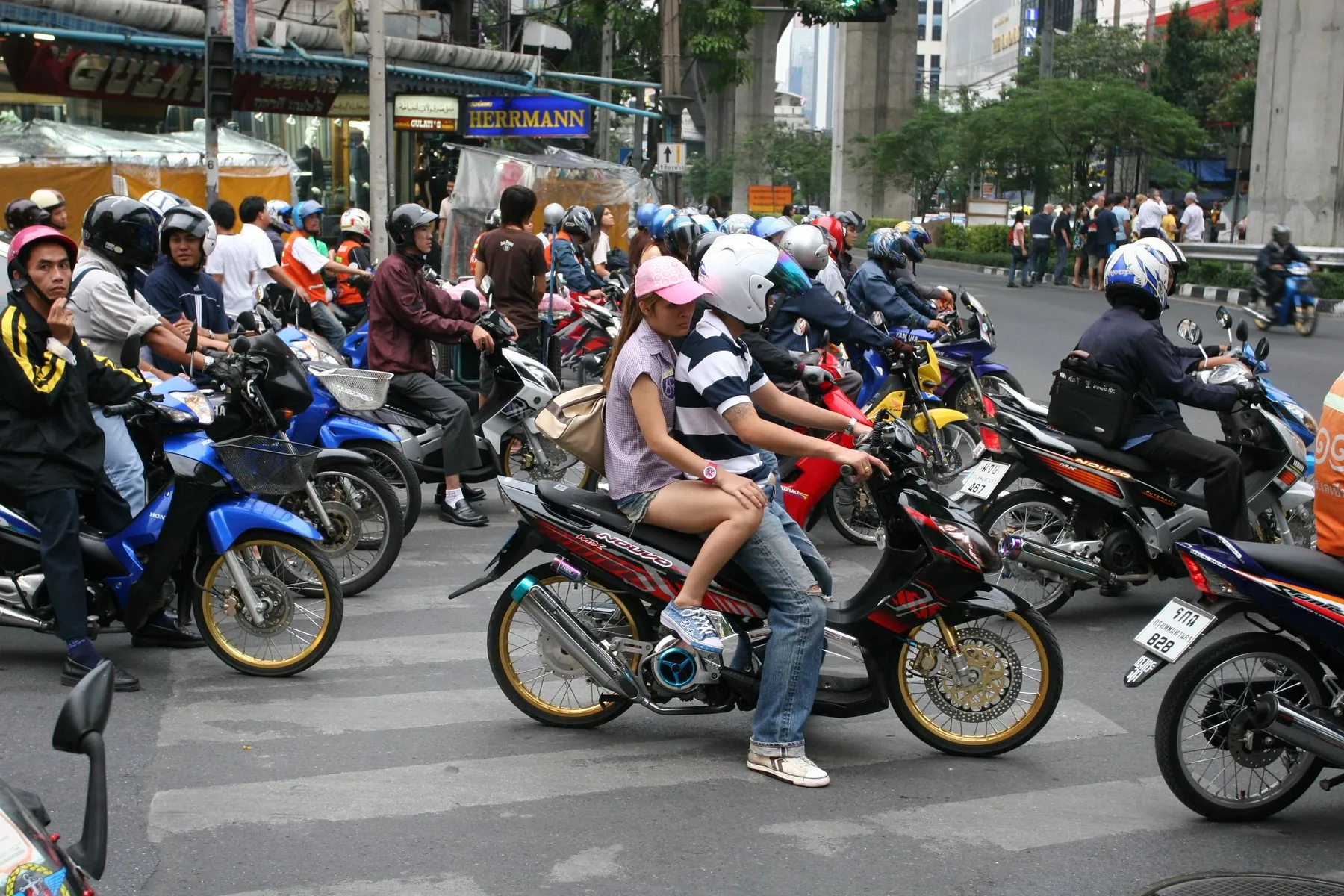 A pack of scooters and small motorbikes waiting at a Bangkok crossing, one carrying a pillion passenger sitting side-saddle