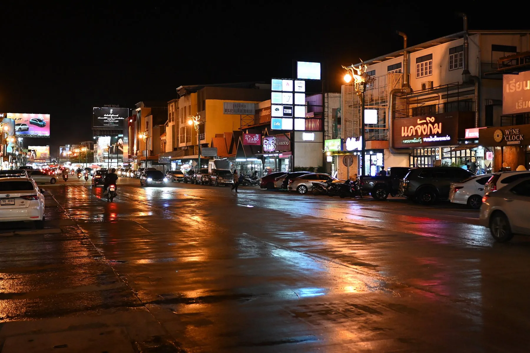 A wide Thai street after dark, the wet tarmac reflecting orange shop signs and the tail lights of passing traffic