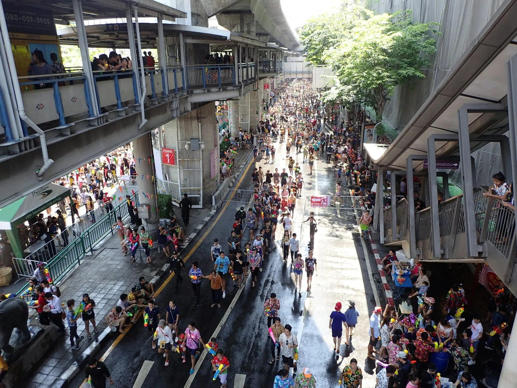 Songkran revellers filling a wet Bangkok street beneath an elevated walkway, seen from above