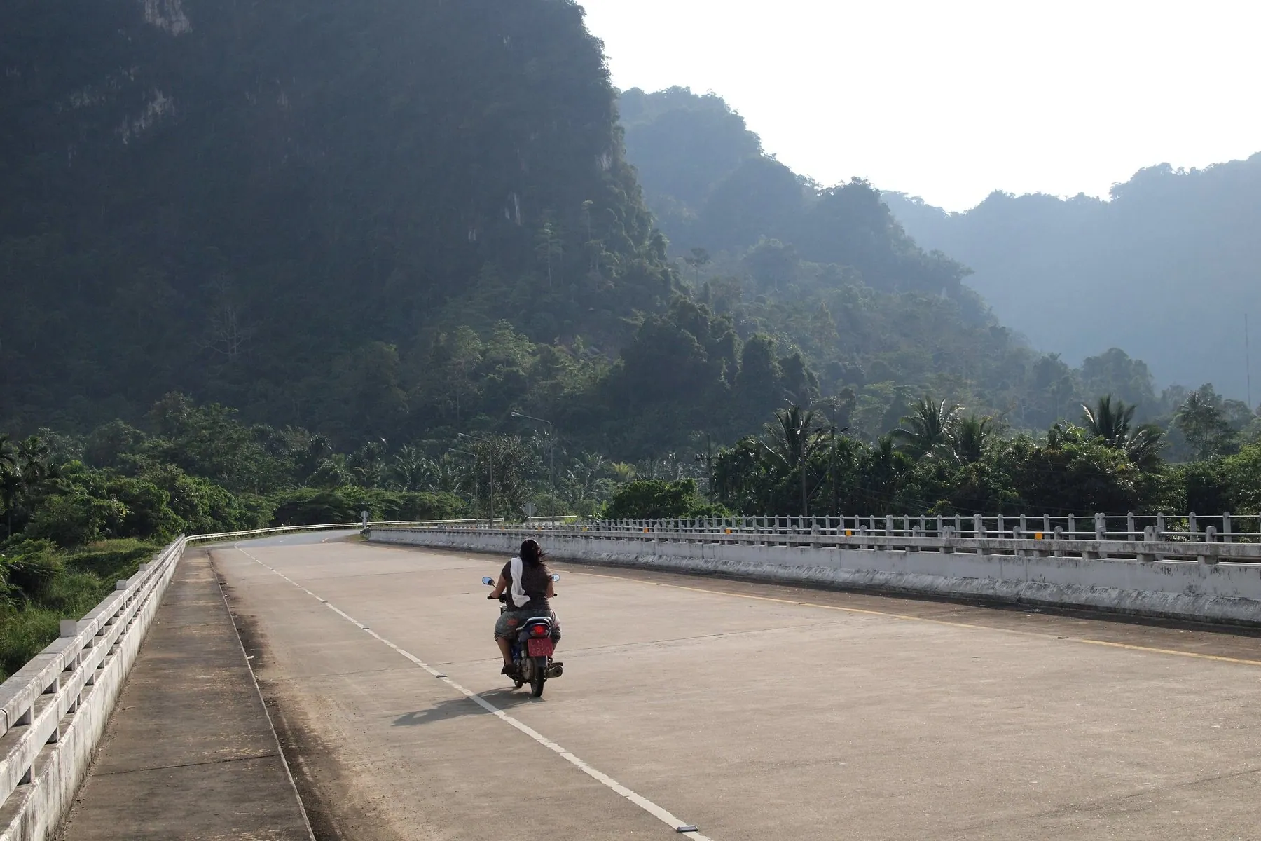 A single motorbike on a long empty causeway running towards limestone hills in Surat Thani