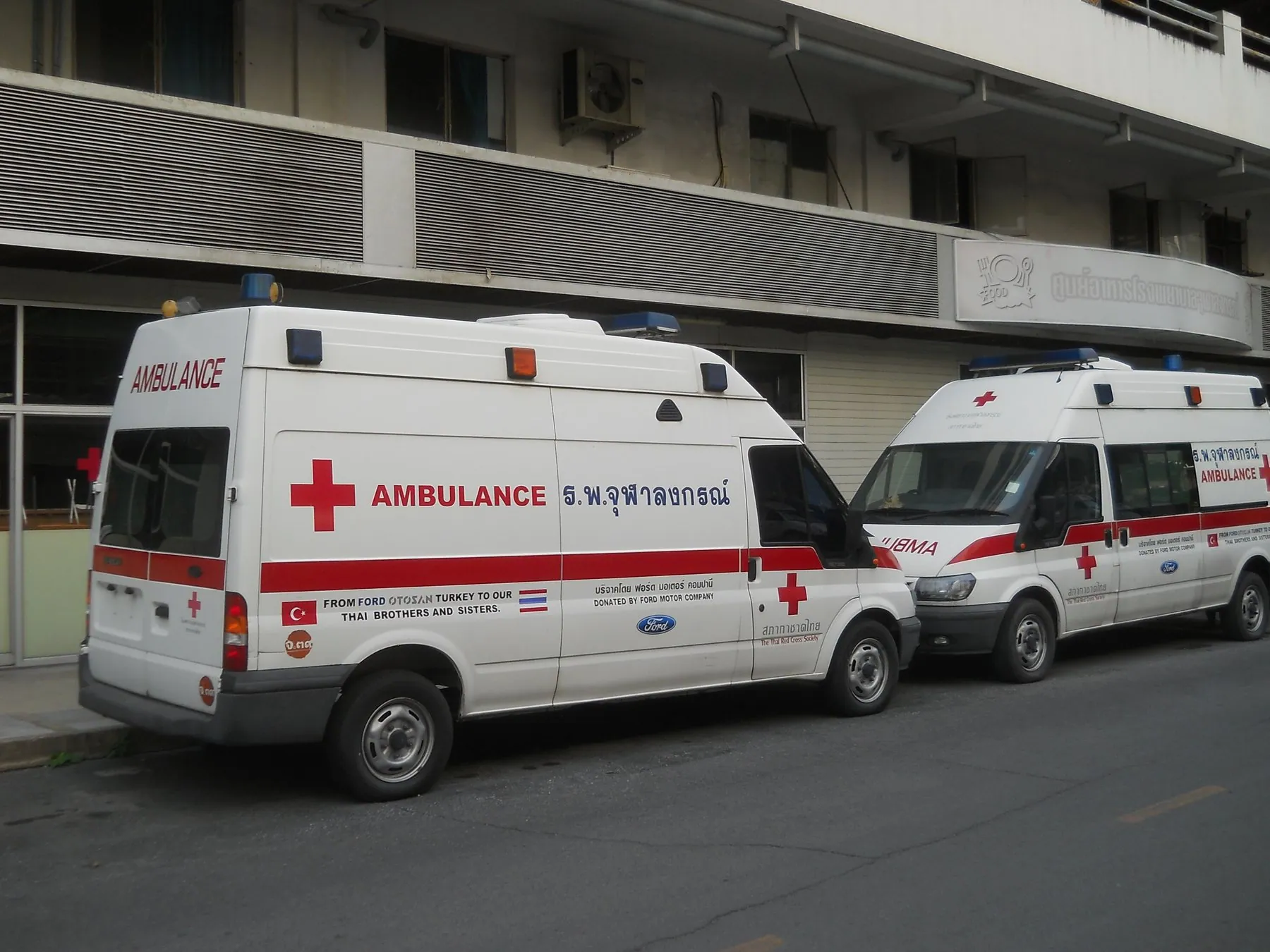 Two Red Cross ambulances parked along a Bangkok street outside a hospital building
