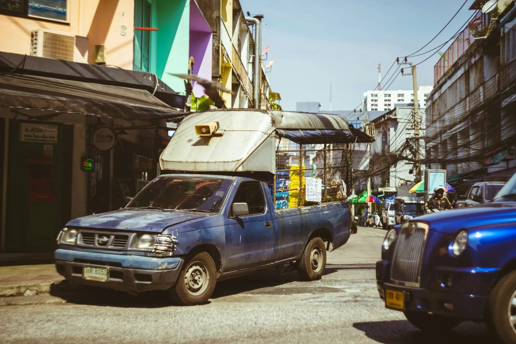 An old blue pickup coming down a narrow Bangkok street lined with shophouses and tangled overhead cables
