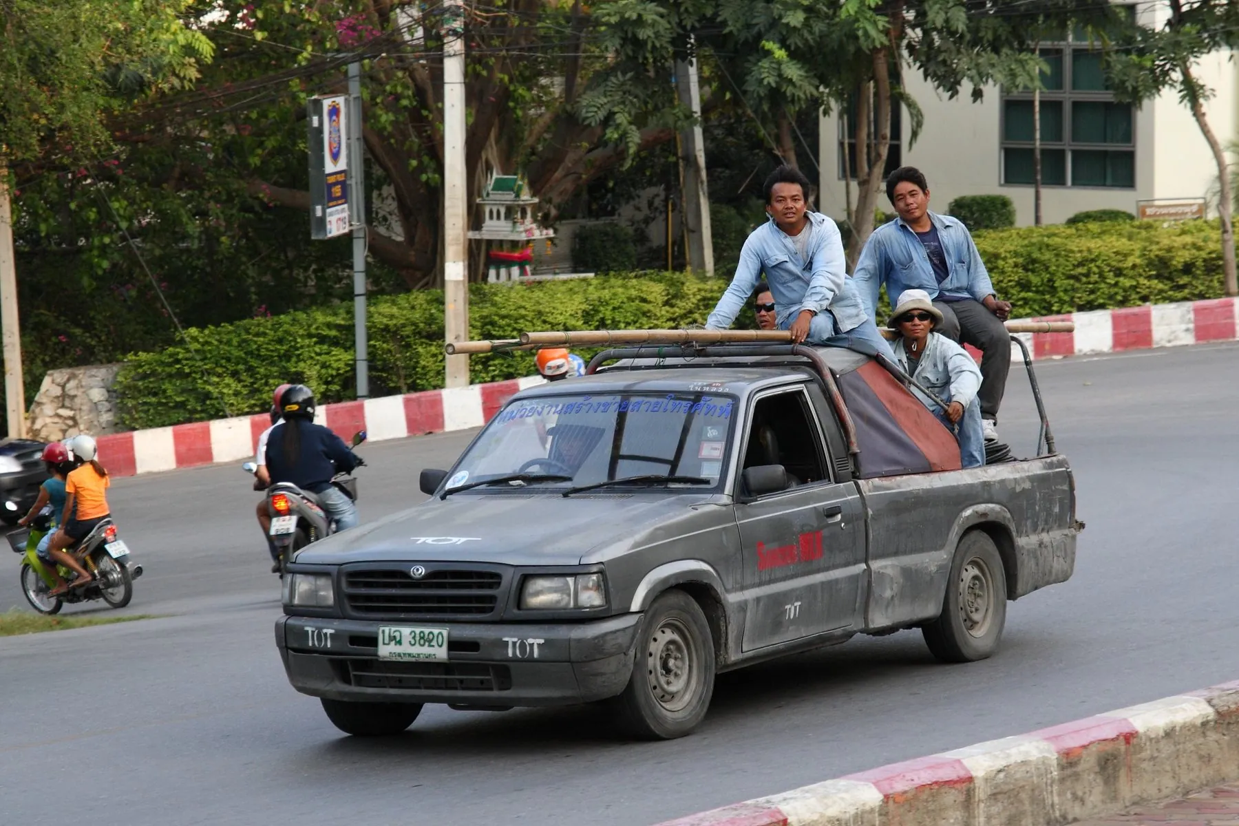 Workers sitting on the sides of an open pickup bed as it is driven along a Thai road with motorbikes alongside