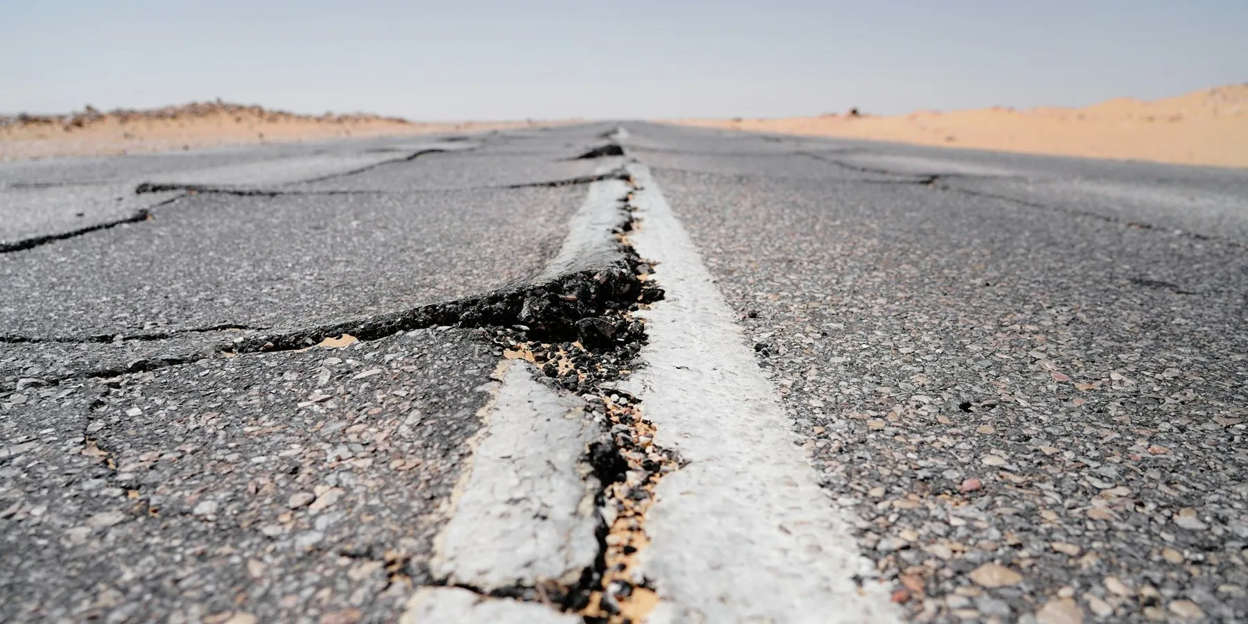 The crumbling edge of an asphalt road at eye level, a long crack running beside the white edge line