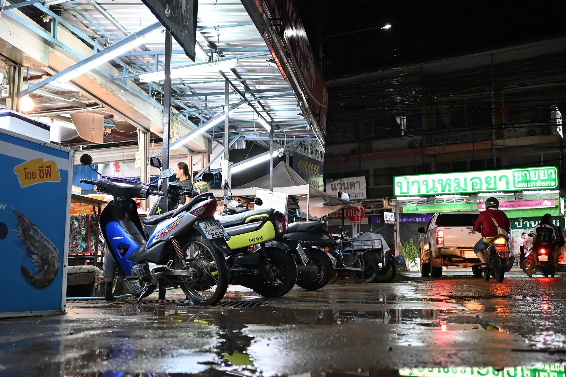 Scooters parked at the kerb of a rain-soaked Thai street while a rider in a poncho passes through the puddles