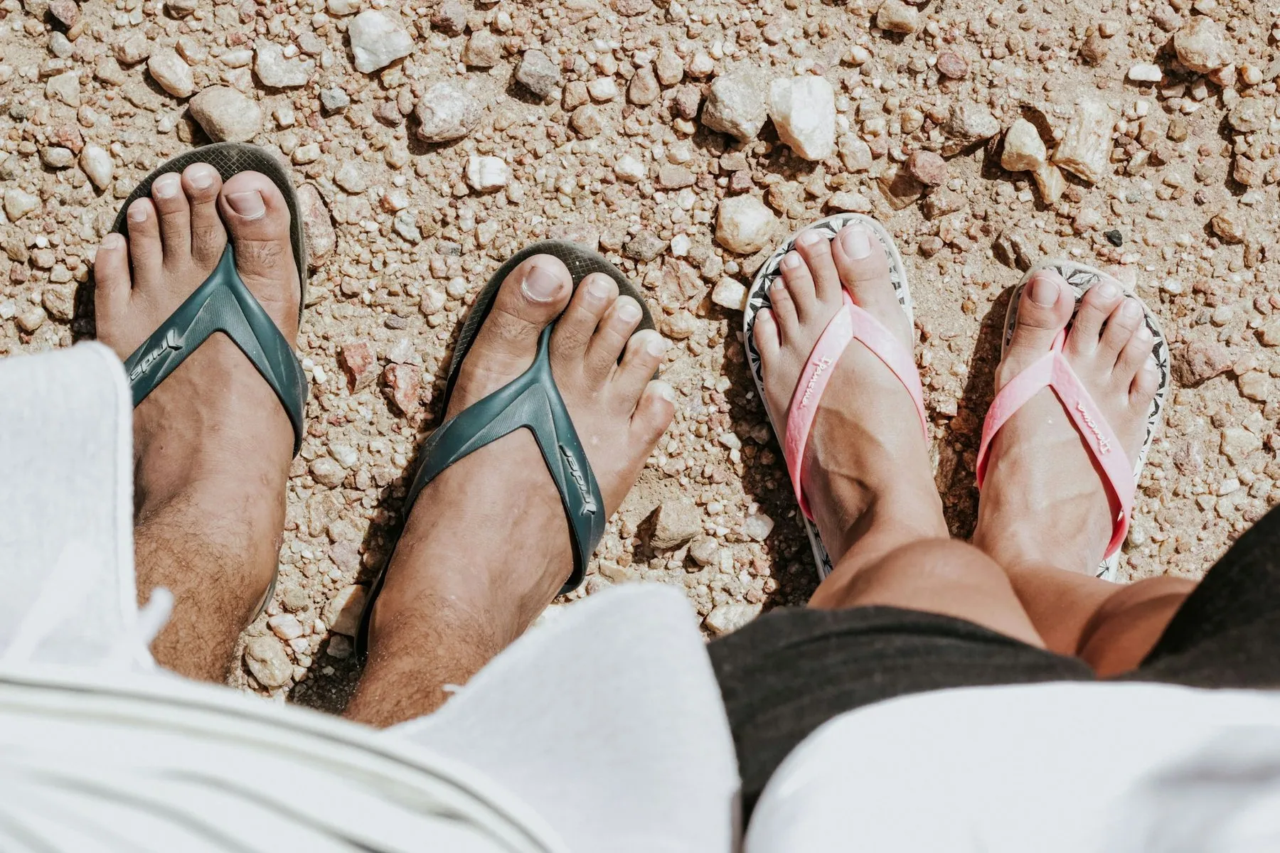 Two pairs of bare feet in rubber flip-flops standing on loose gravel