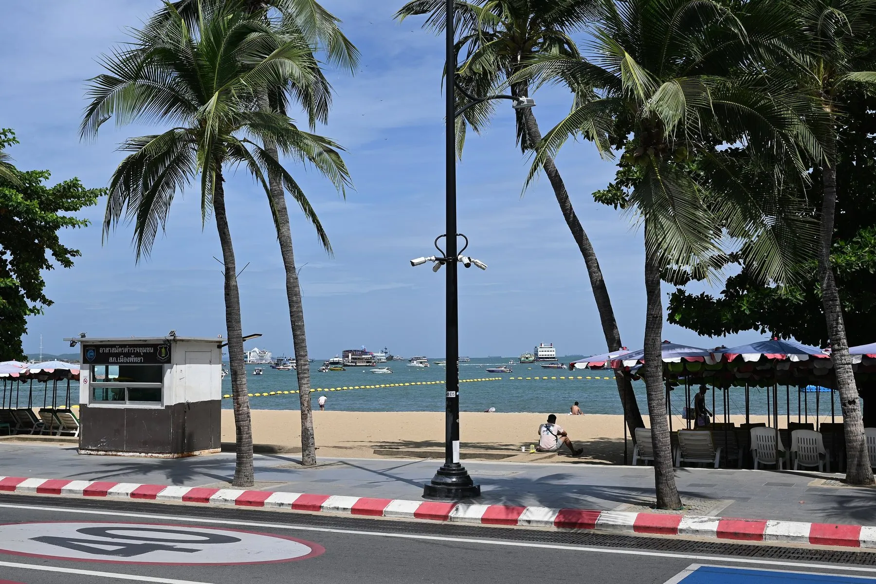 A quiet coastal road with a red and white painted kerb running past palm trees and open sand to the sea