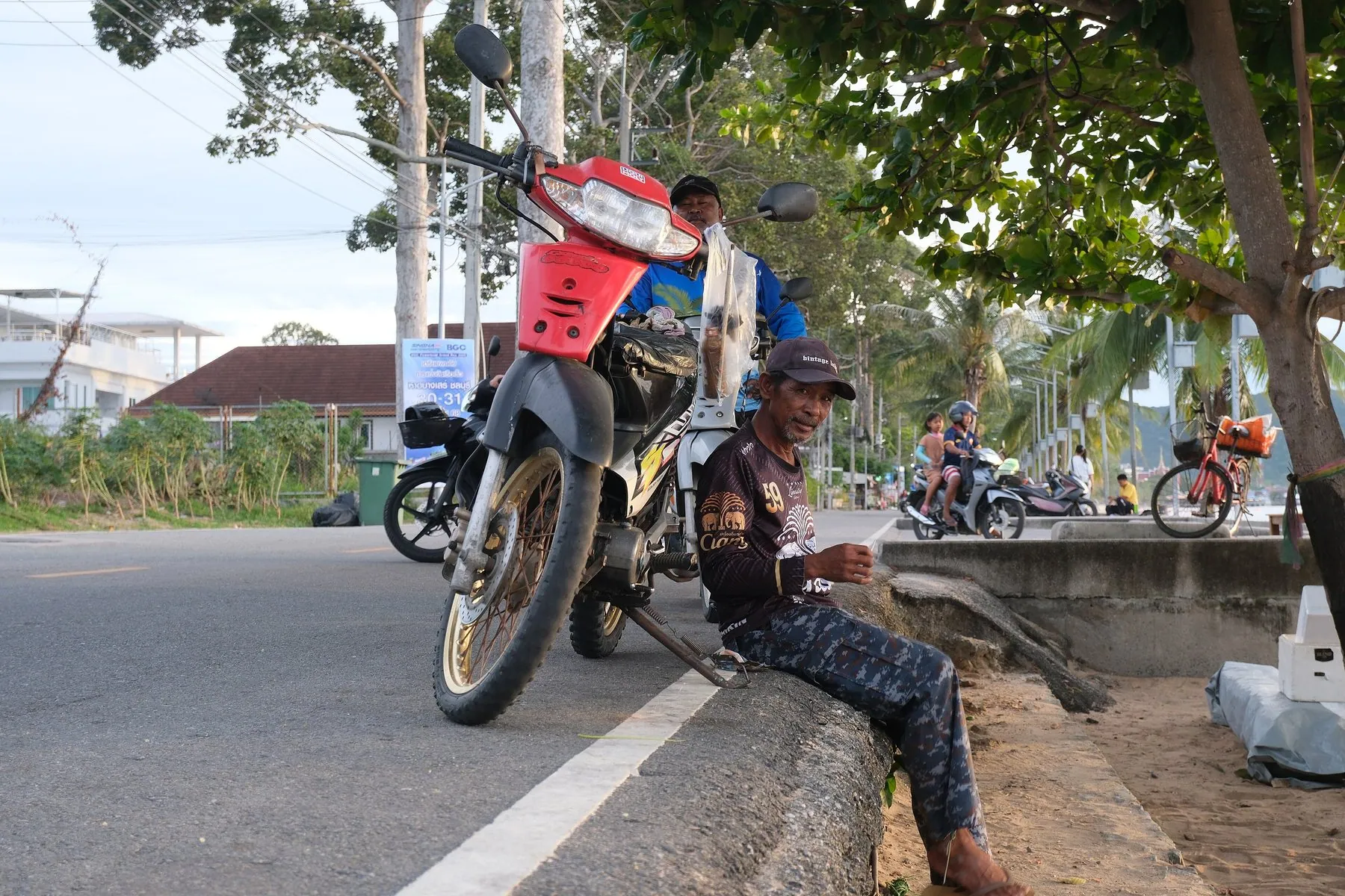 A man resting on a kerb in the shade of a roadside tree beside his parked red motorbike, palms and more bikes further down the road