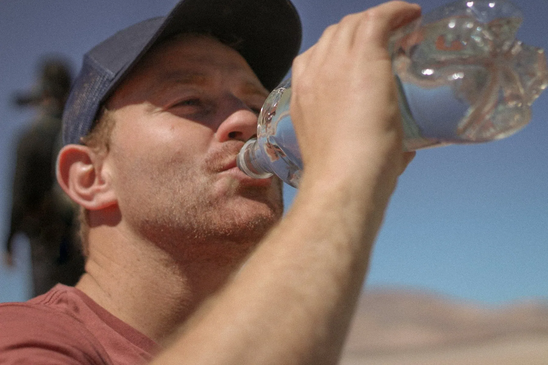 A man in a cap tipping back a plastic water bottle in bright sunlight