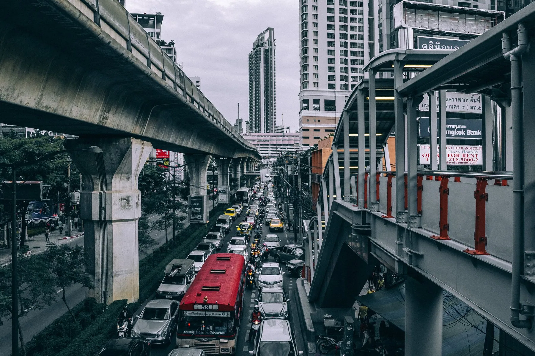 Bangkok traffic queued nose to tail beneath the concrete deck of the skytrain, seen from a footbridge
