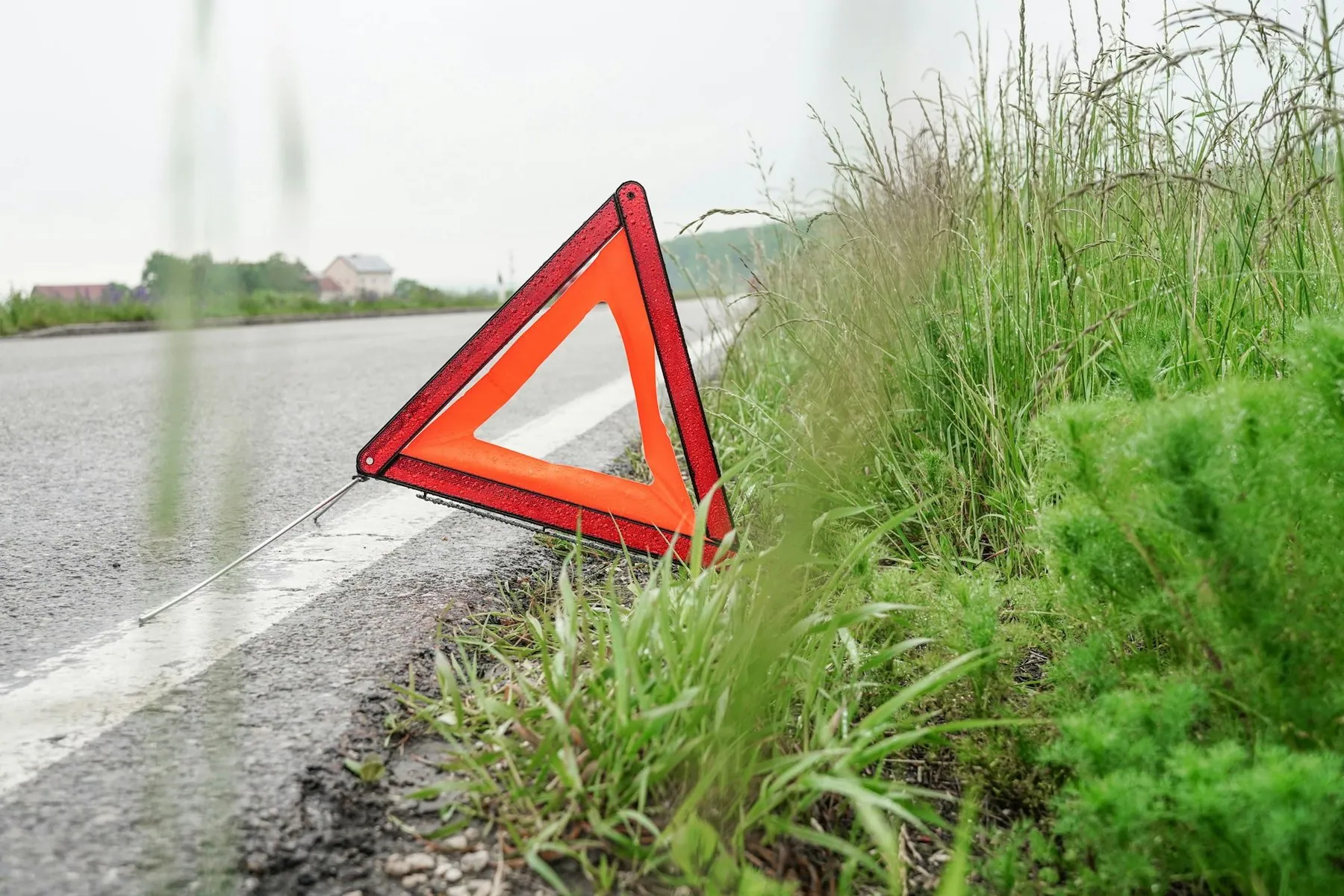 A red reflective warning triangle standing on the grass verge at the edge of a wet road