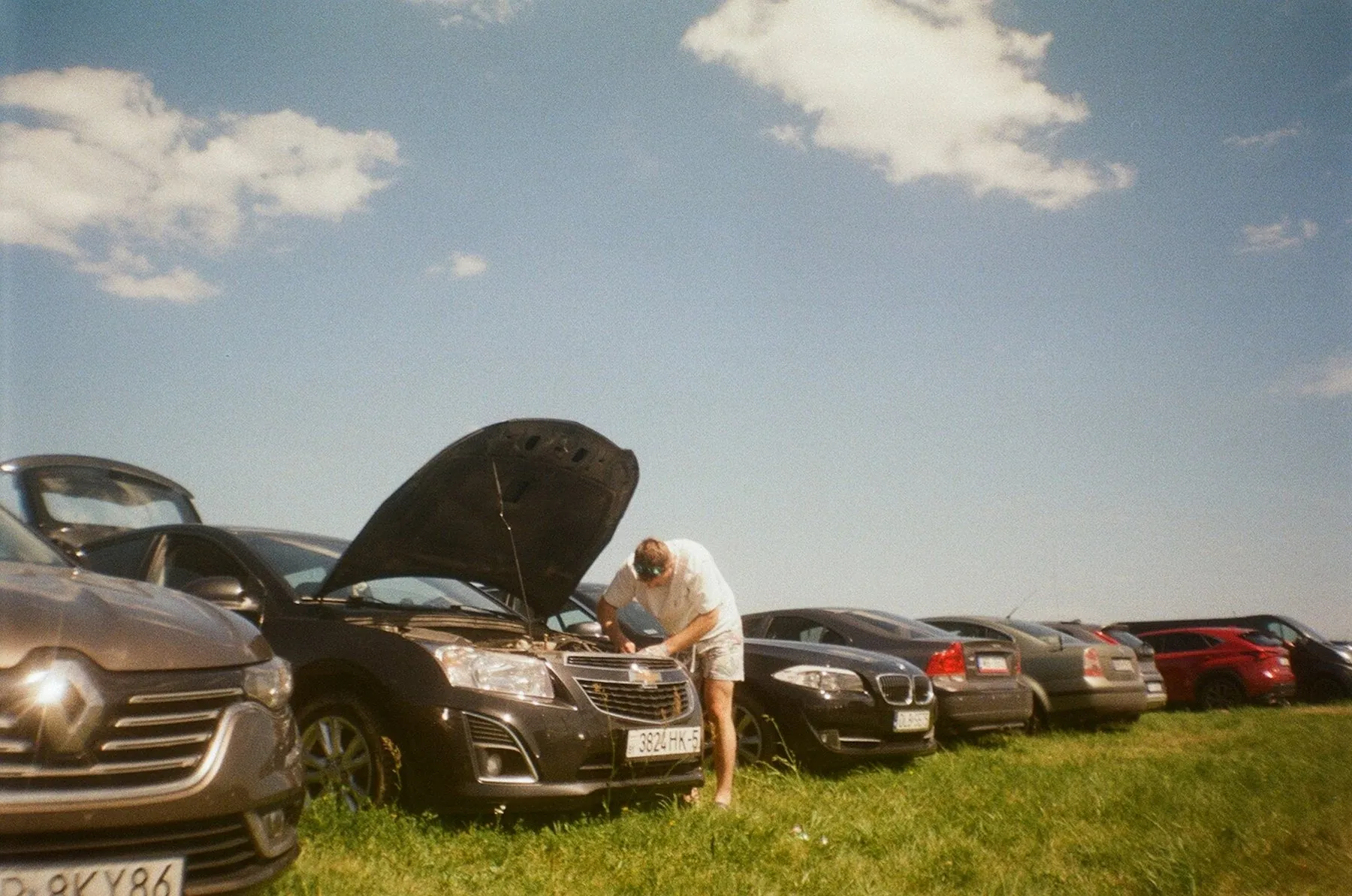 A man leaning over the open bonnet of a car parked on rough grass at the edge of a road