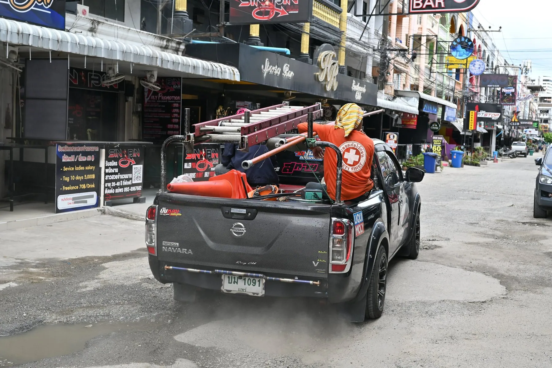 Rescue volunteers in orange riding in the open back of a pickup stacked with equipment as it pulls away down a street of Thai shopfronts