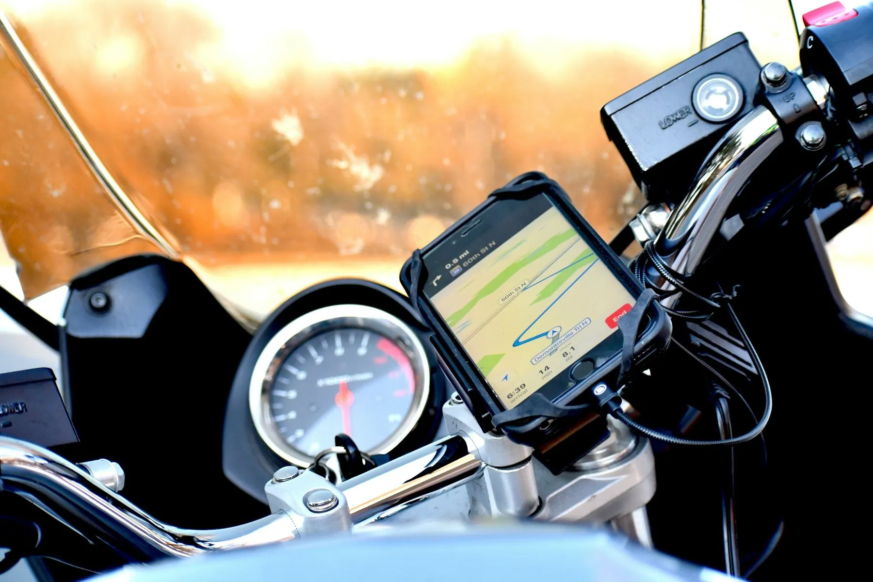 A smartphone showing a navigation map clamped to motorbike handlebars beside the speedometer, rain on the windscreen